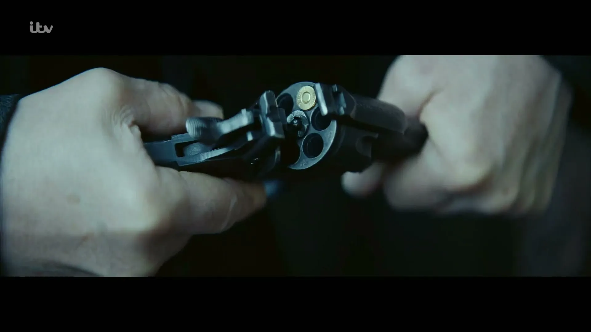 A man's hands are seen in a close-up shot, loading a single bullet into a revolver in a dimly lit interior.