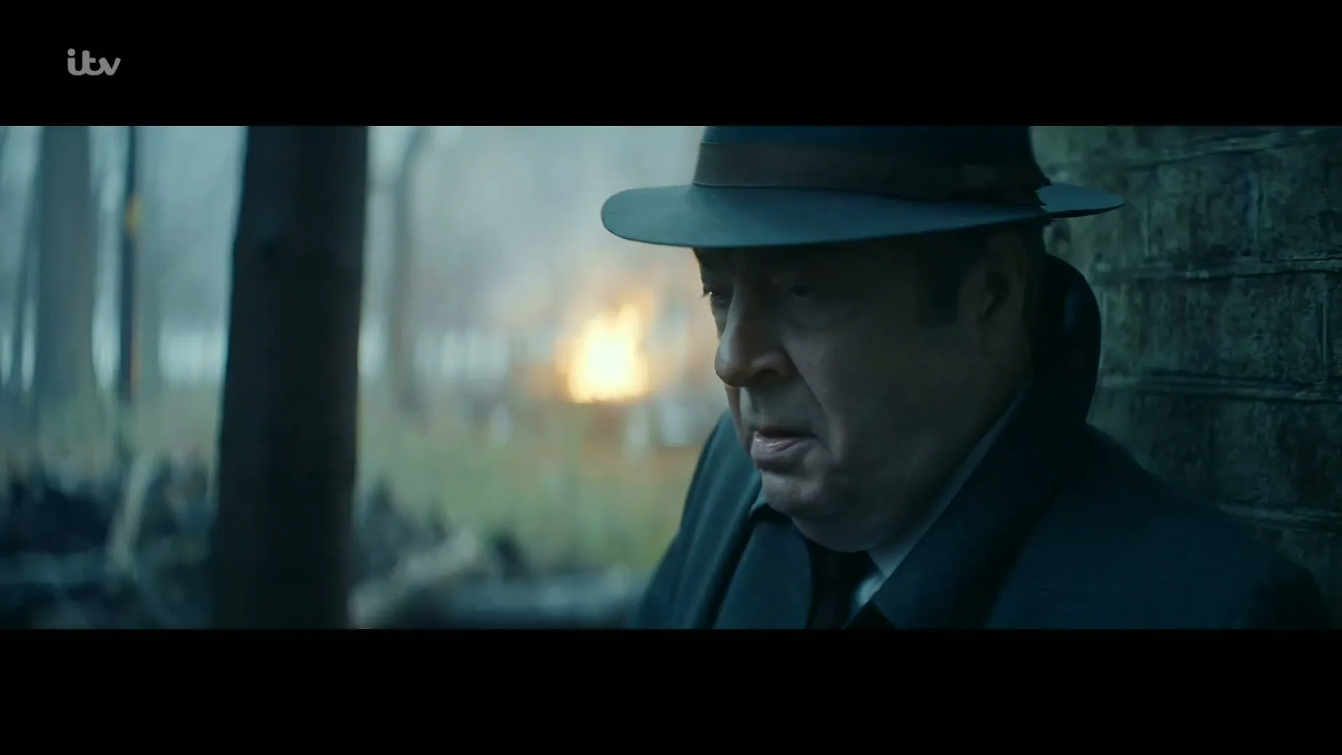 A man in a dark coat and fedora hat looks distressed, leaning against a brick wall with a fire burning in the background.