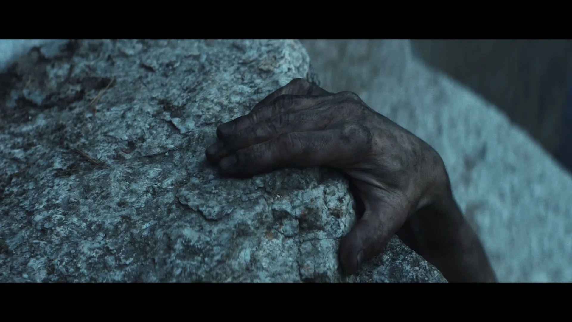 A dirty, weathered hand grips a rough, grey rock, struggling to hold on in a close-up shot.