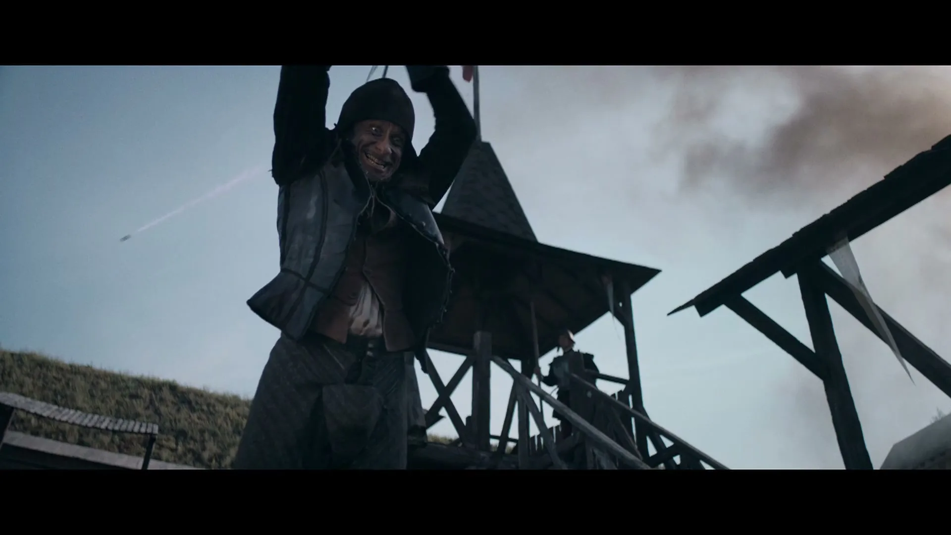 A male pirate with a scarred face and dark clothing swings a sword, grinning maniacally on a wooden structure under a cloudy sky.