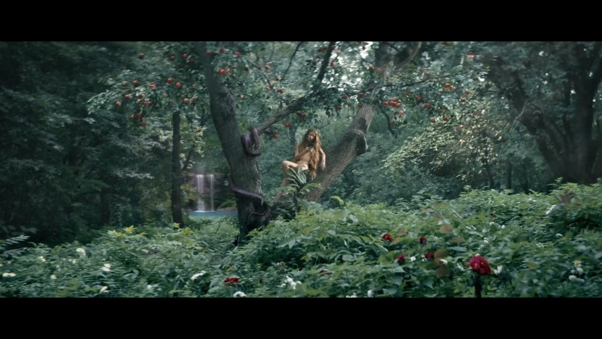 A long-haired woman, resembling Eve, sits naked in a tree with red apples and a snake, in a lush, green forest with a waterfall in the background.