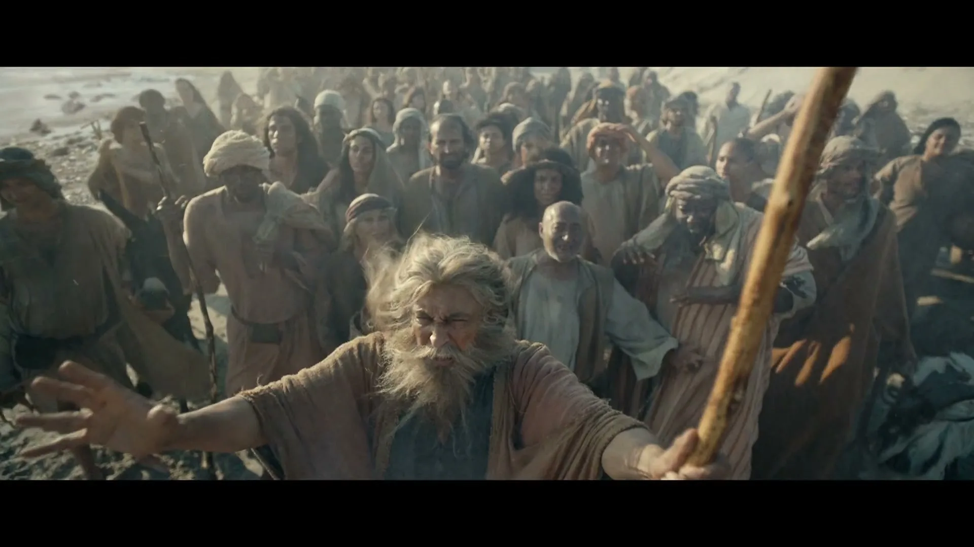 An old man with a long white beard and a staff leads a large crowd of people through a desert landscape, looking determined.
