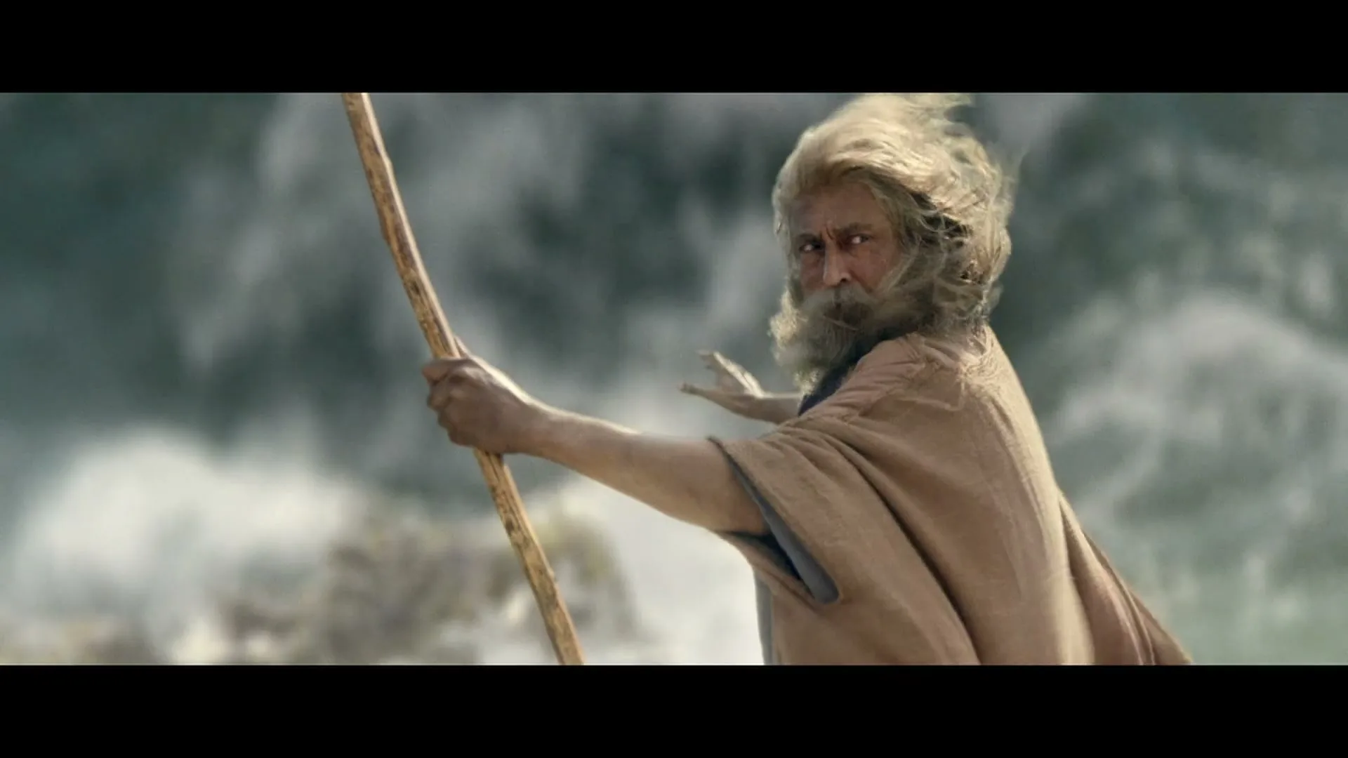 A man with a long white beard and hair, dressed in a brown robe, holds a staff and turns to look at the camera with a surprised expression, as water crashes behind him.