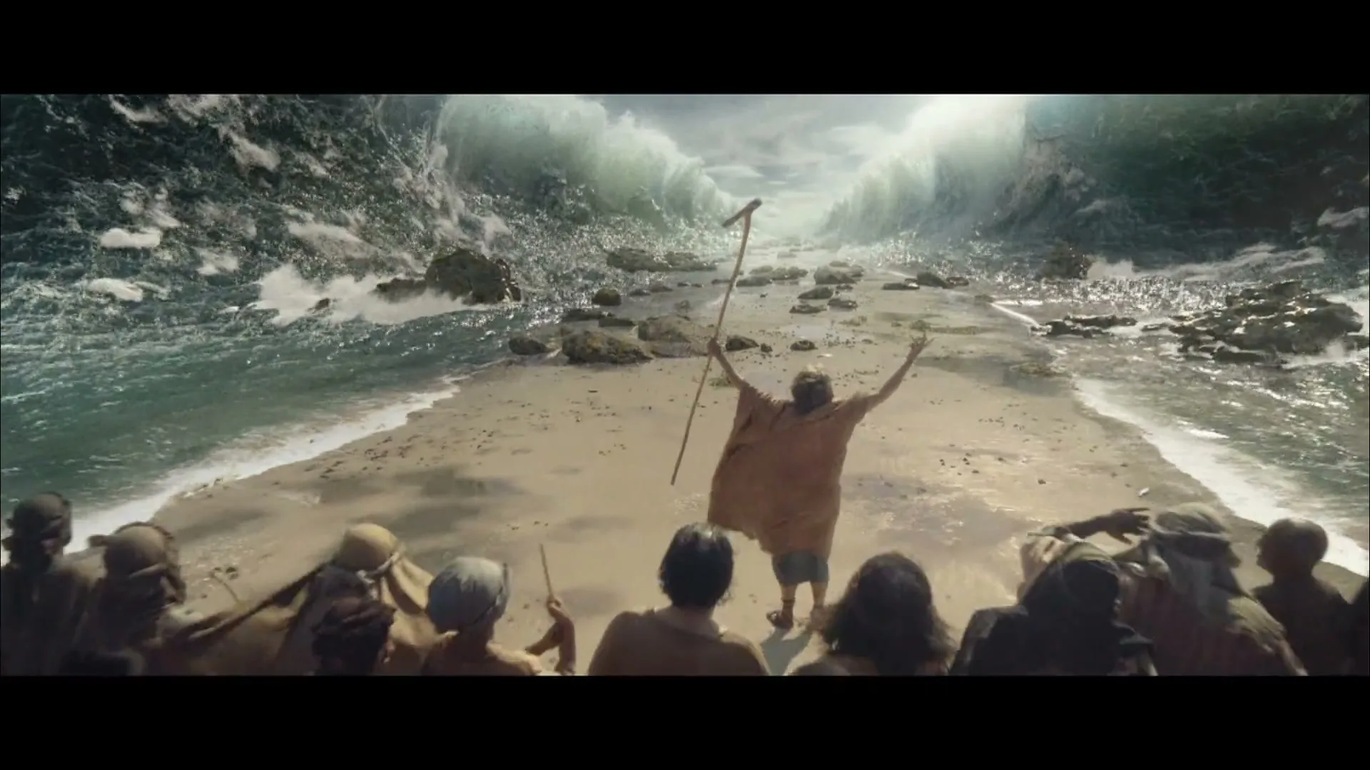 A man in a brown robe with a staff stands on a sandy path between two towering walls of water, with a crowd of people gathering behind him.
