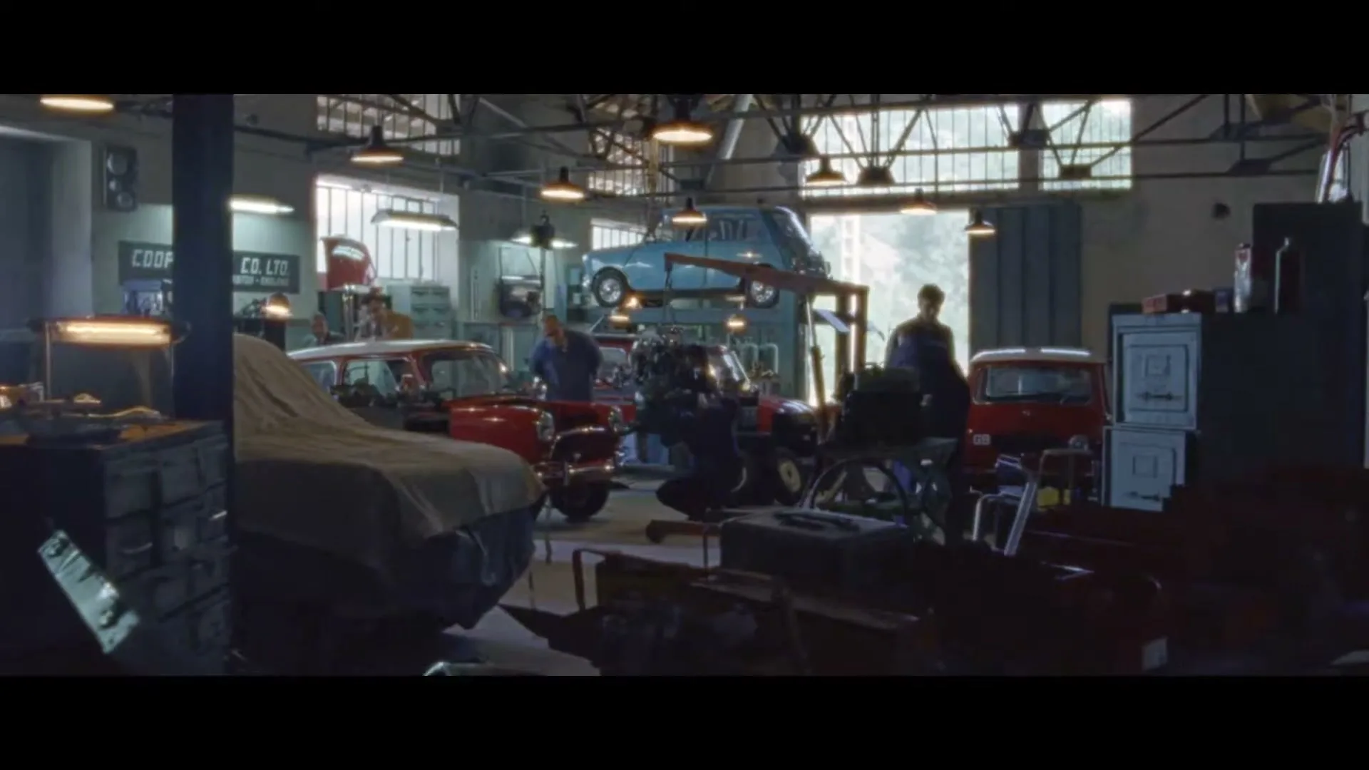 A wide shot of a busy car garage with several mechanics working on classic Mini cars, one of which is elevated on a lift.