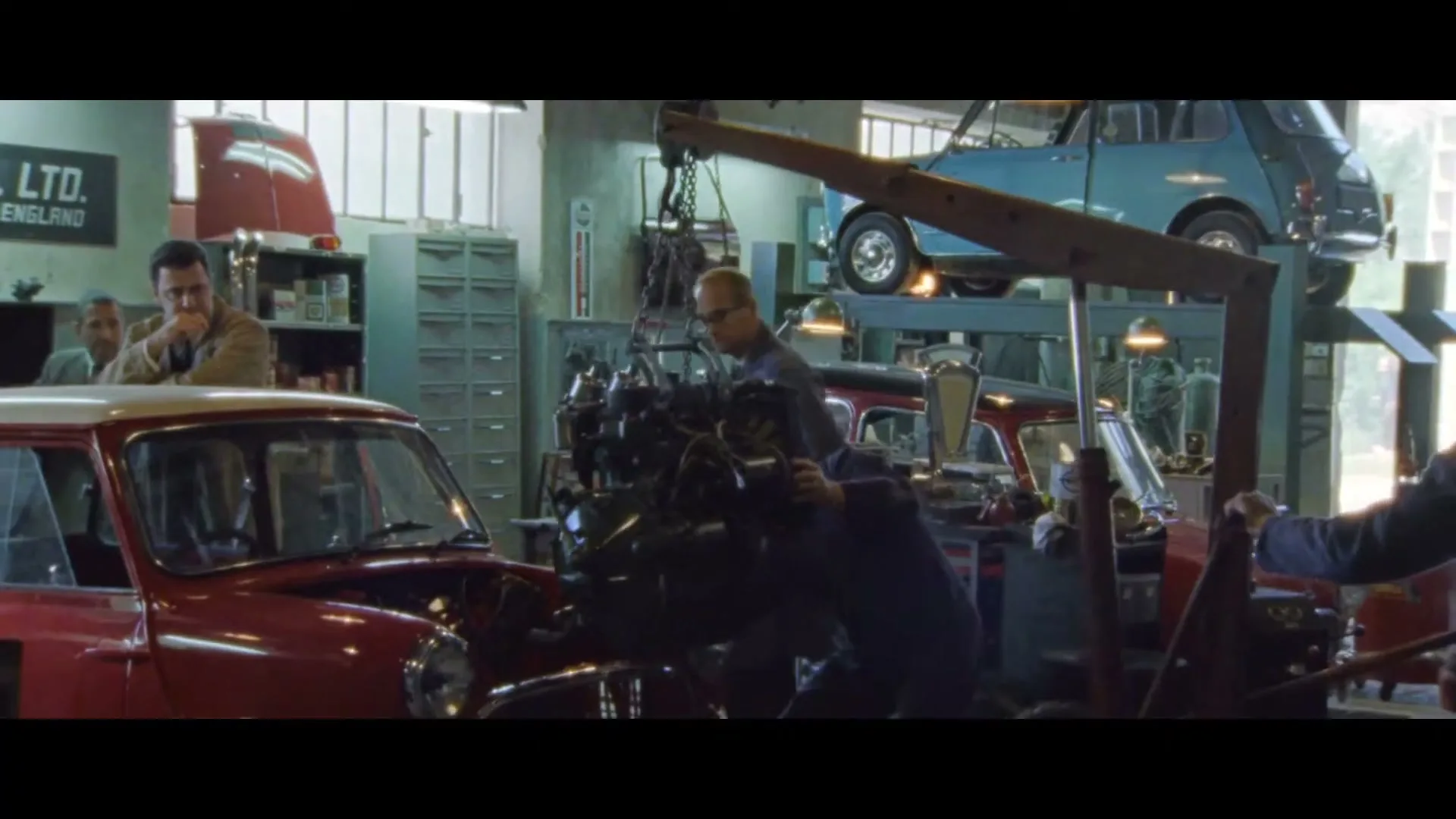 A group of mechanics work on classic Mini cars in a busy garage, with one man lowering an engine into a red car.