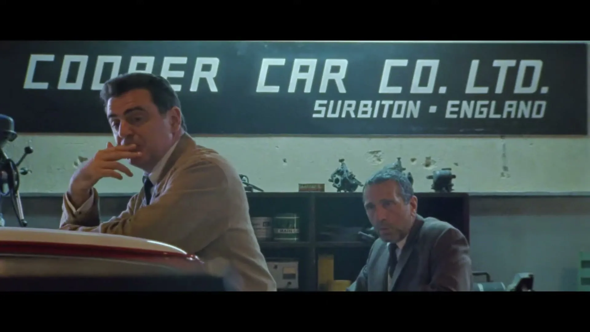 Two men in suits are in a garage with a "Cooper Car Co. Ltd." sign in the background, one man is leaning on a car, the other is sitting at a desk.