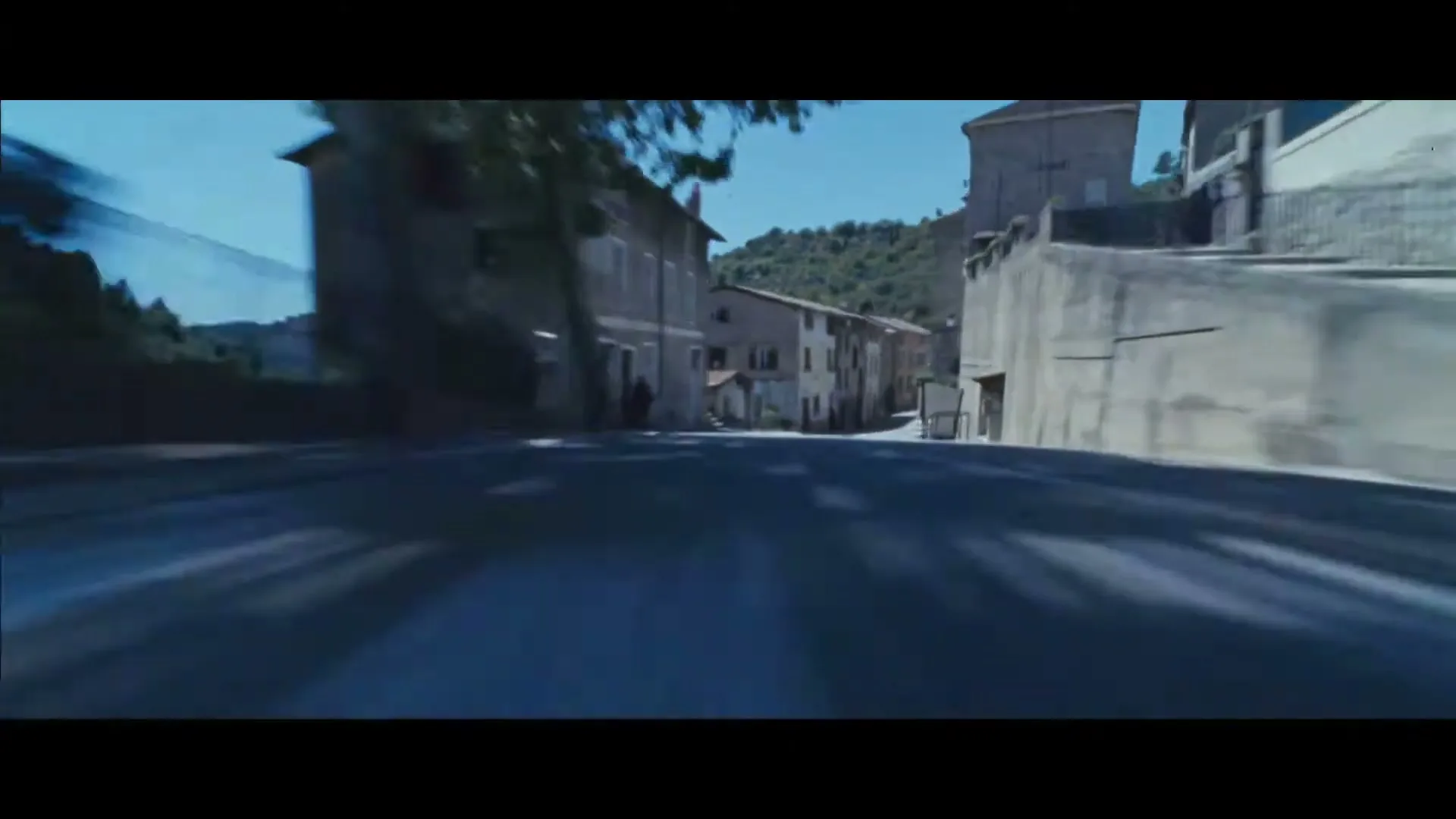 A low-angle POV shot from a car speeding through a narrow, sunlit street in a European village.