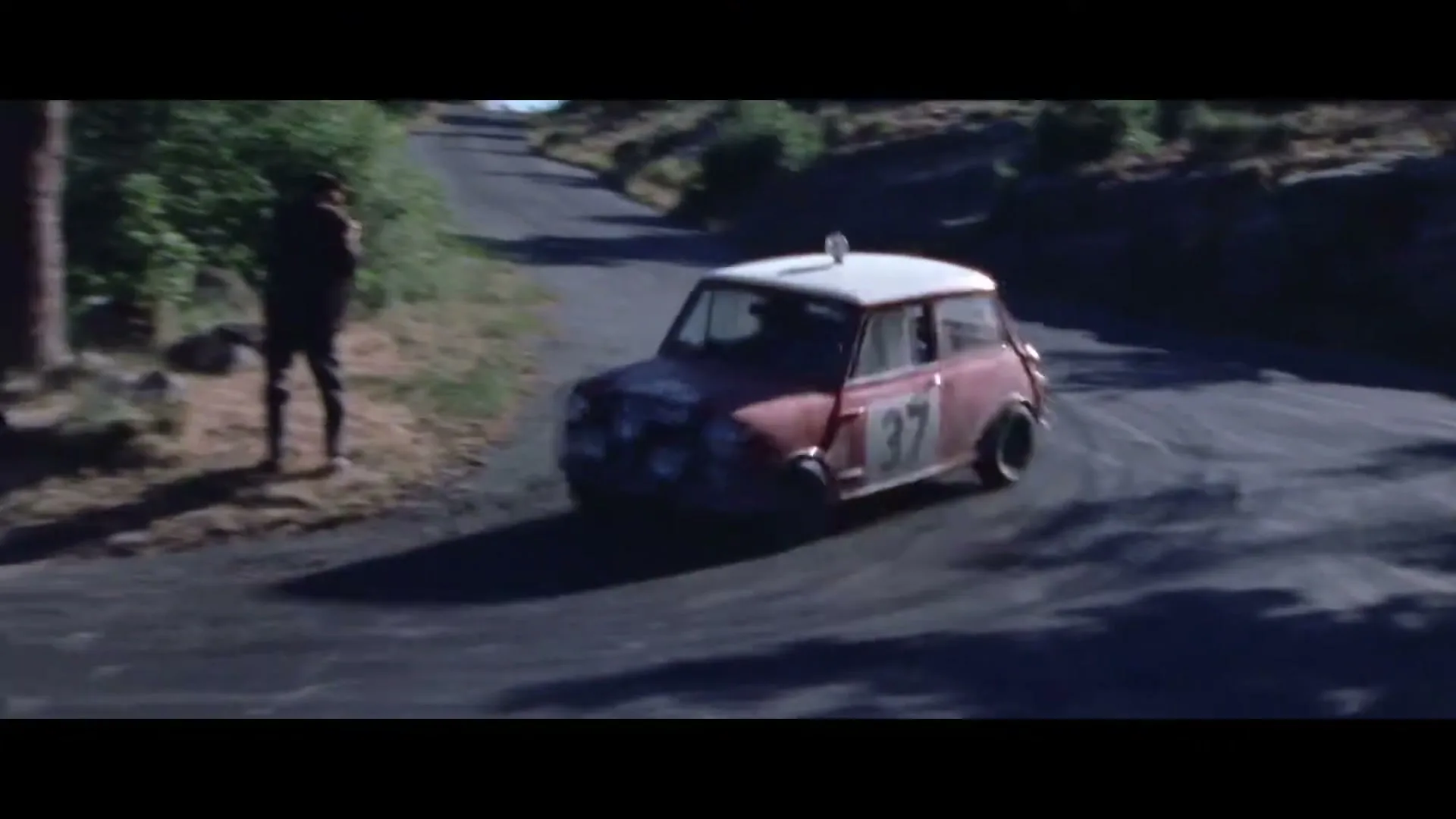 A red Mini Cooper rally car with number 37 on the side drifts around a corner on a winding mountain road, with a person watching from the side.