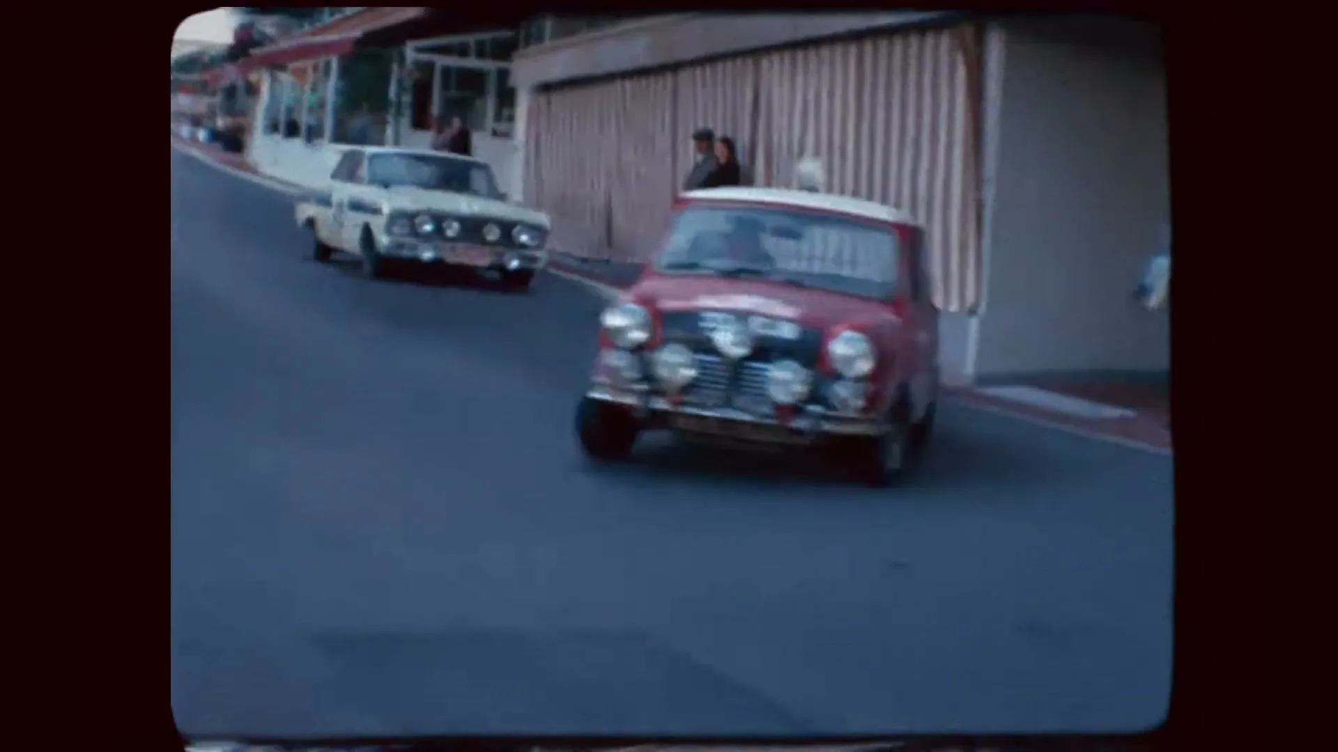 A red Mini Cooper with the number 37 on its side races up a street, passing a white car and spectators on the sidewalk.