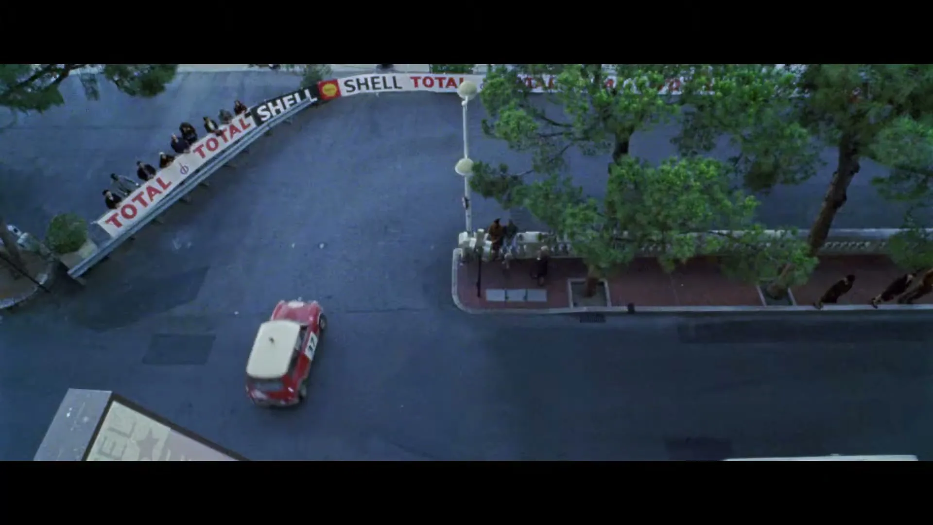 A red and white Mini Cooper races around a corner on a city street, with spectators and trees visible.
