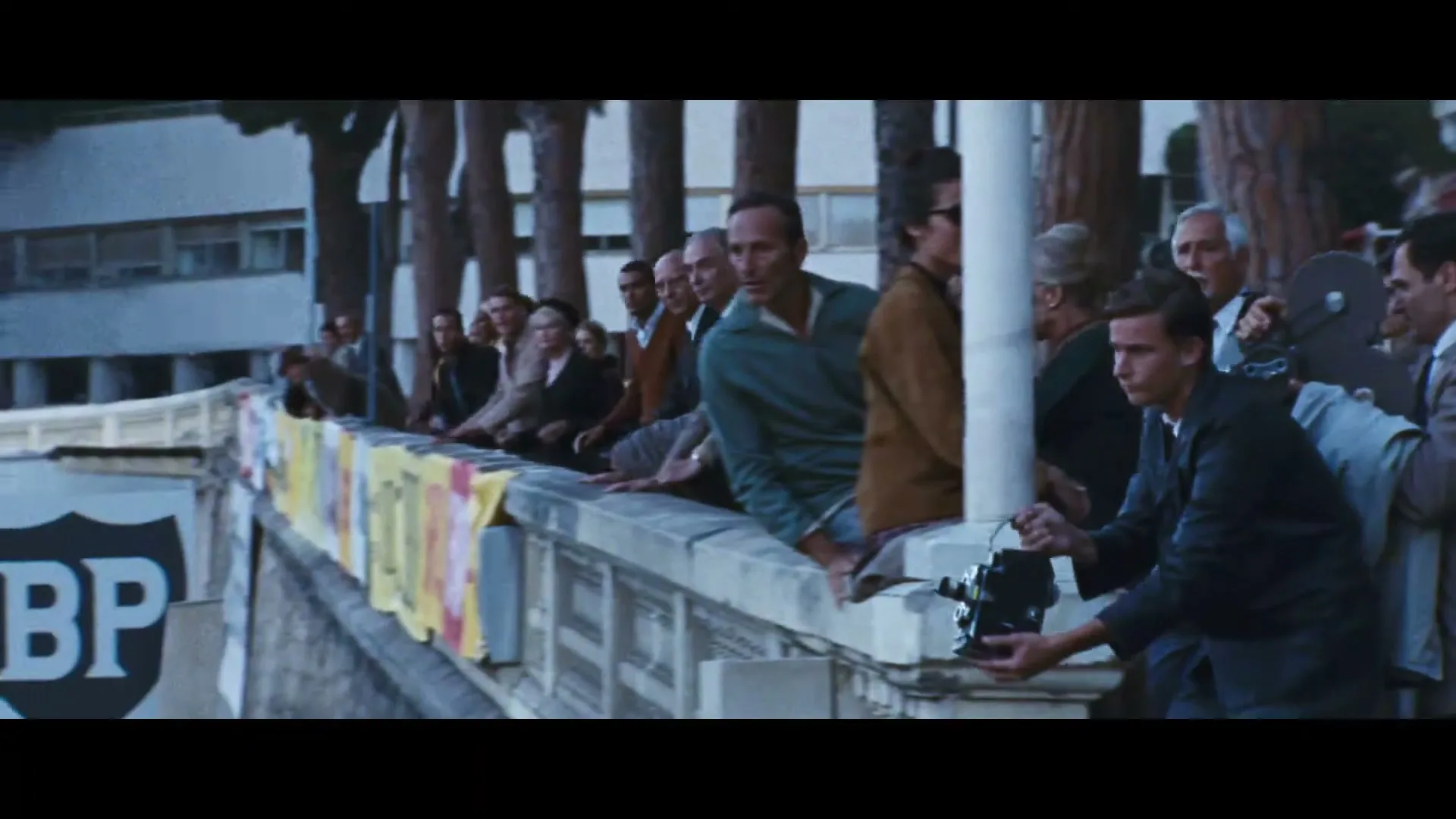 A crowd of people, including a man with a camera, watch a red car speed by on a race track during the day.