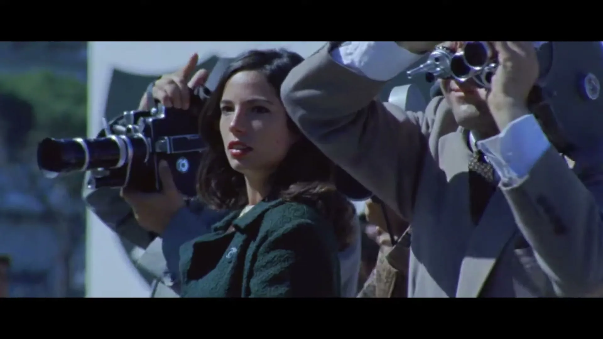 A young woman in a green coat stands among photographers with vintage cameras, looking intently to her right in an outdoor setting.