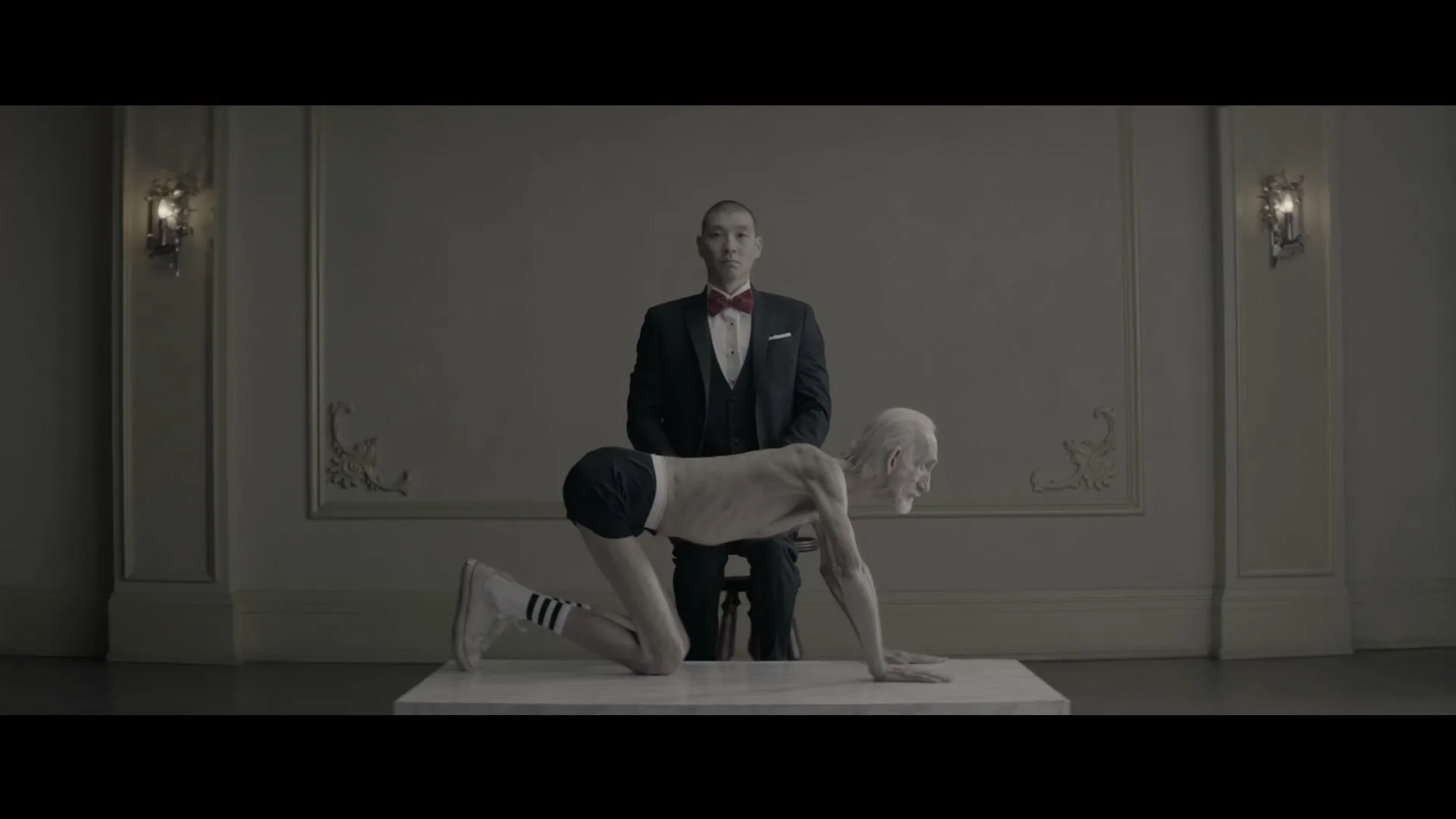 A man in a tuxedo sits behind an elderly, emaciated man on all fours, in a formal room.