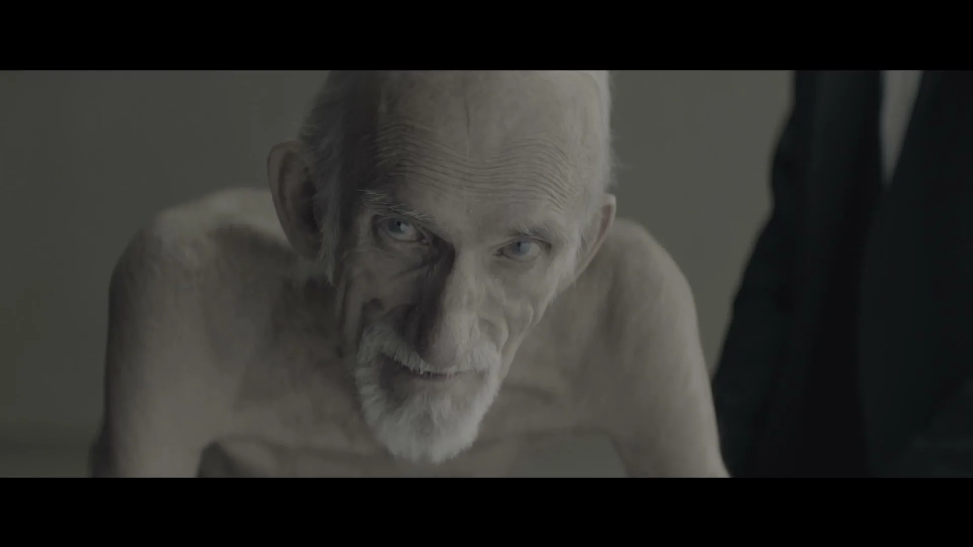 An old, emaciated man with a white beard and blue eyes looks directly at the camera with a slight smile, in a dimly lit room.