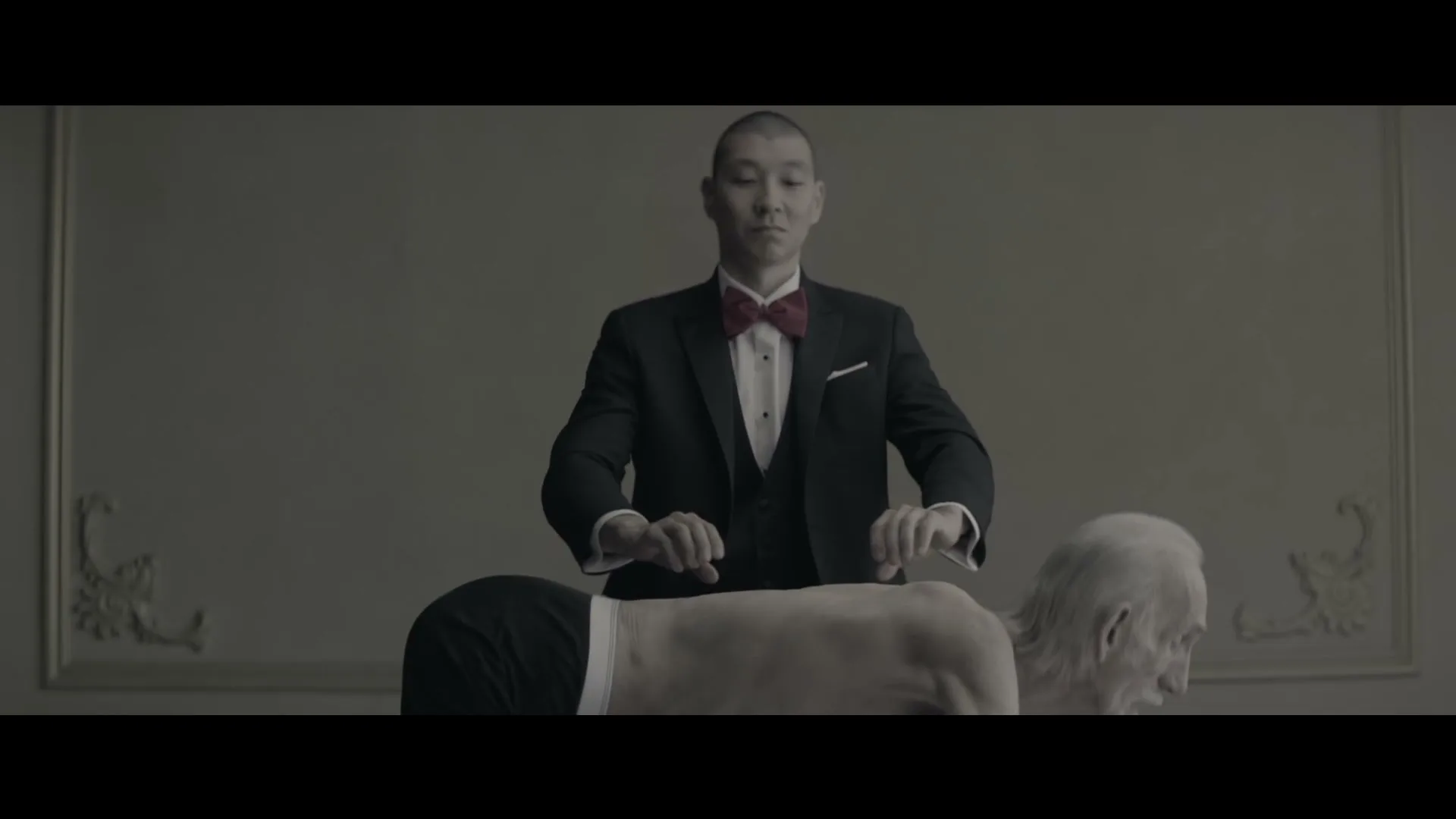 A man in a tuxedo plays a piano on the back of an old man, who is bent over in a formal room.