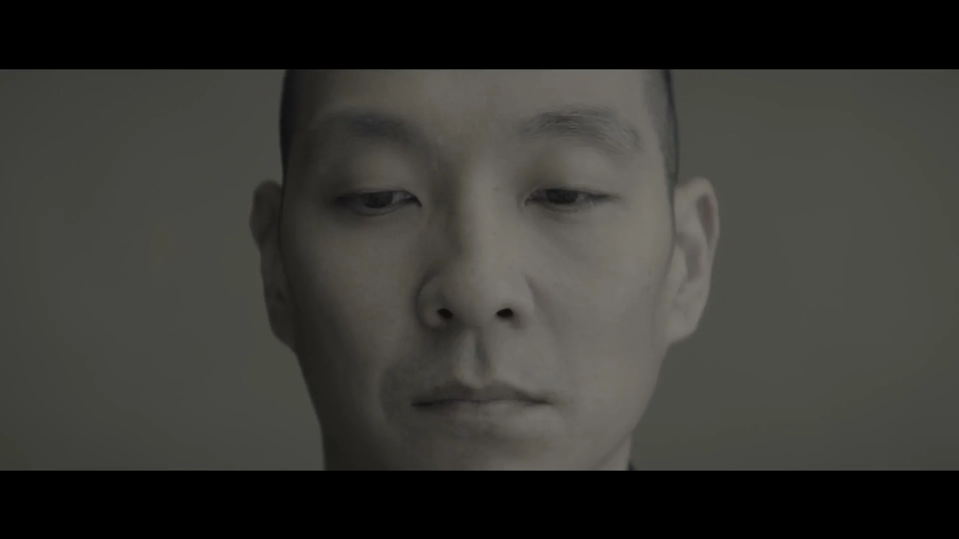 A man with a shaved head looks down, then slowly raises his eyes to look directly at the camera in a studio.