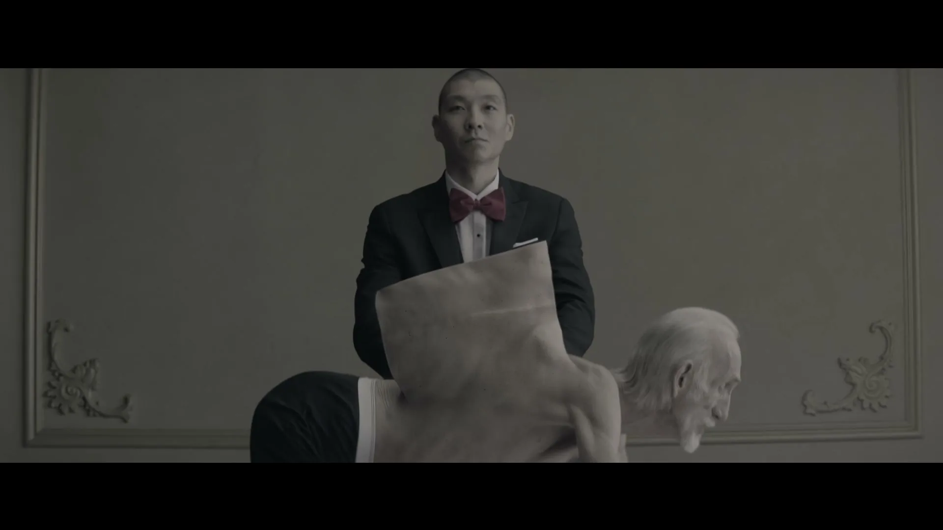 A man in a tuxedo stands behind an older man bent over, with a piece of paper covering his back, in a dimly lit room.