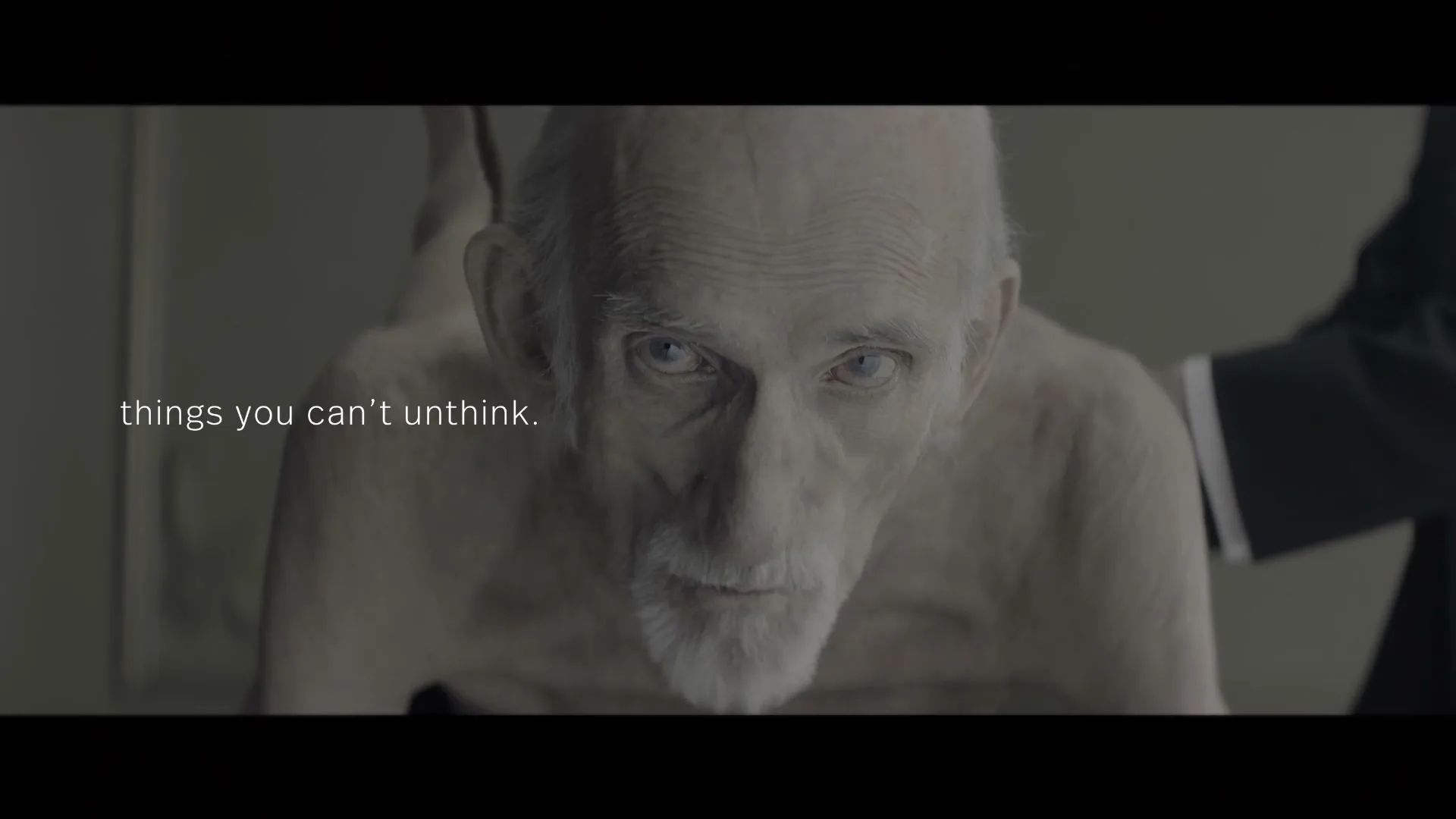 A pale, elderly man with a white beard and blue eyes looks directly at the camera, with a dark-suited arm visible behind him.