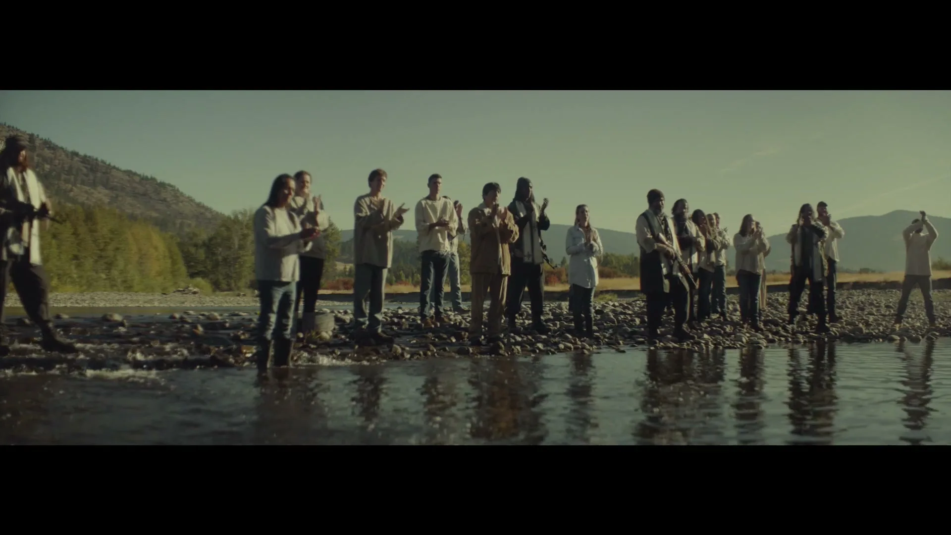 A group of people, dressed in simple, rustic clothing, stand in a shallow river, clapping their hands in unison under a clear blue sky.