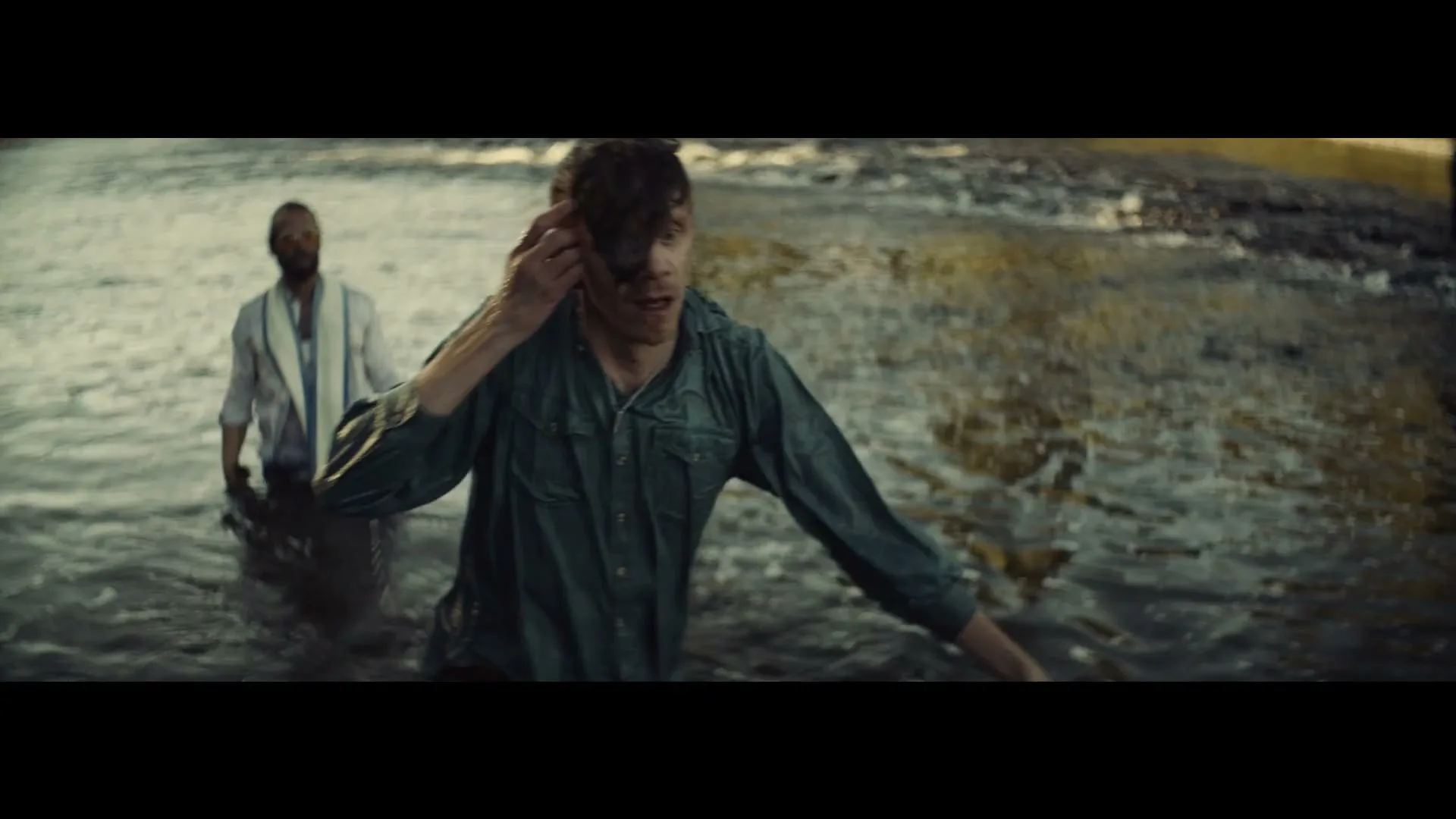 A man in a wet green shirt emerges from a river, wiping water from his face, while another man in a white shirt stands behind him in the water.