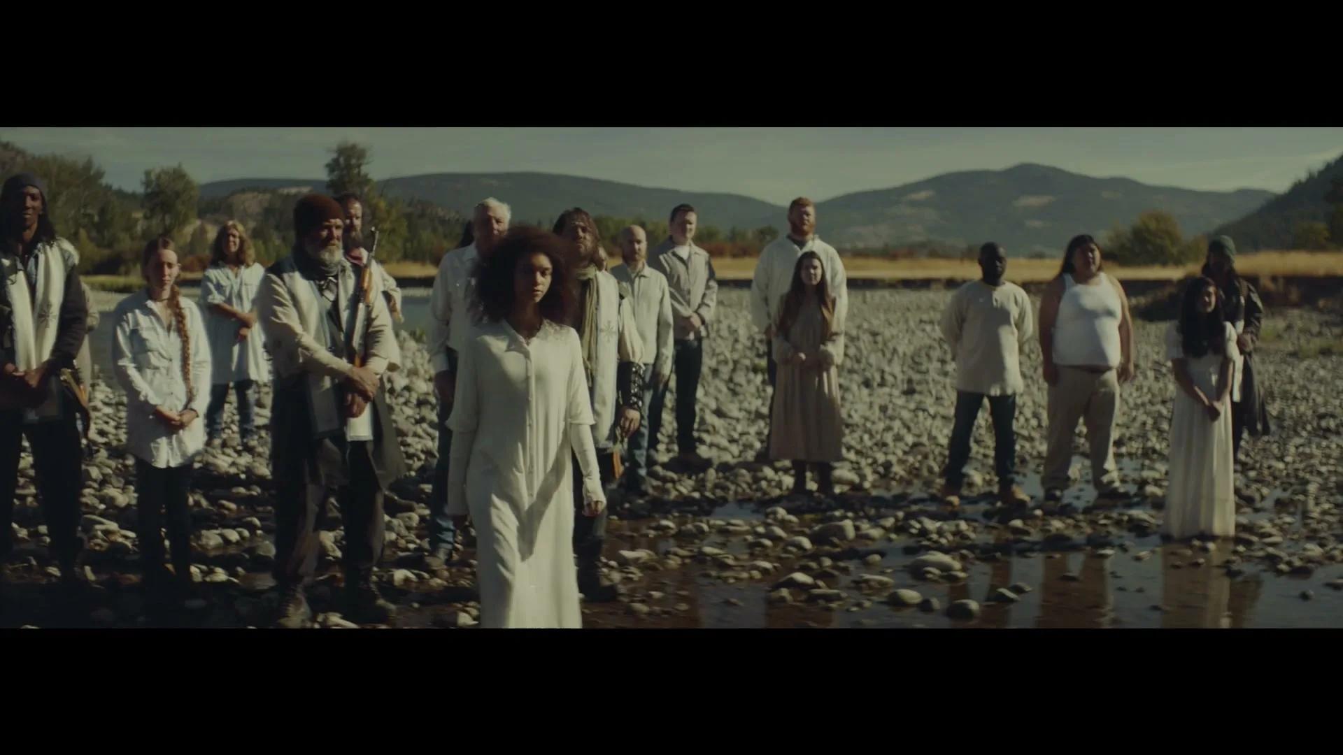 A diverse group of people, some in white robes, stand in a shallow riverbed with mountains in the background on a sunny day.