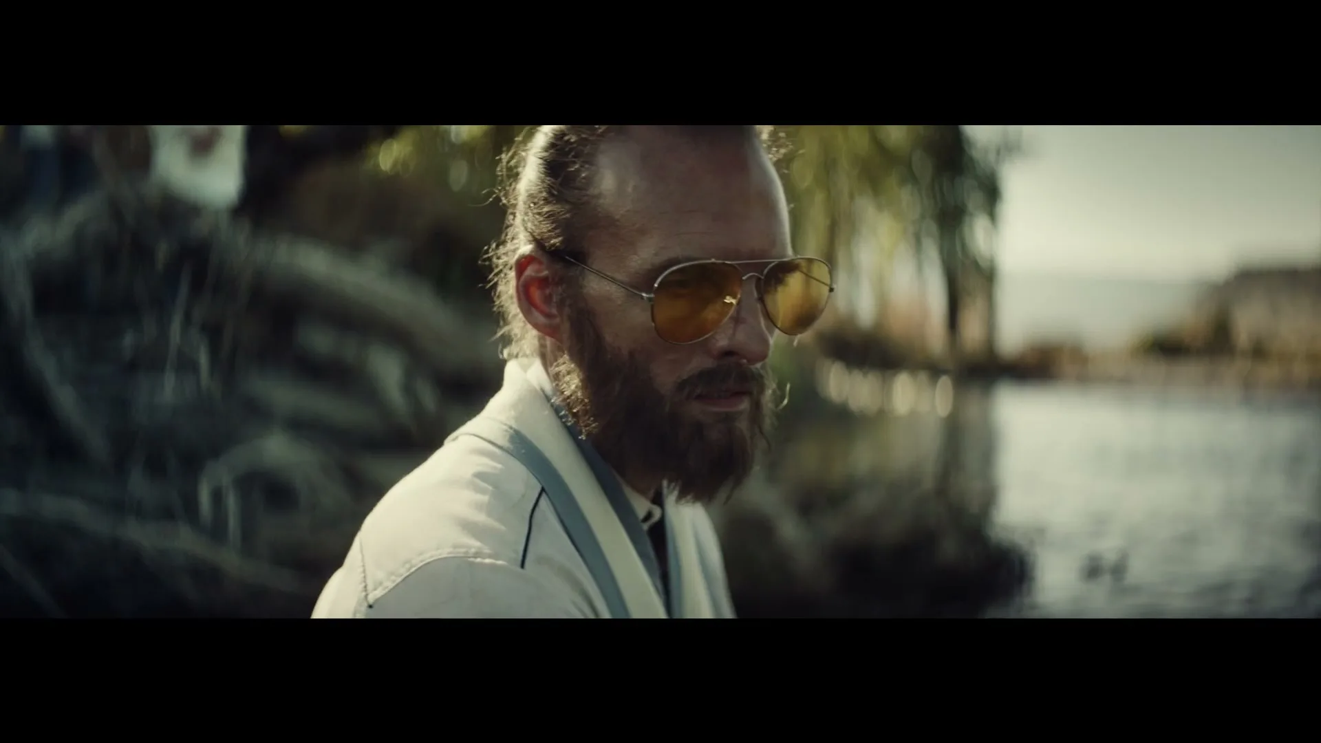 A bearded man with sunglasses and a ponytail looks down, sitting by a body of water in a sunny outdoor setting.