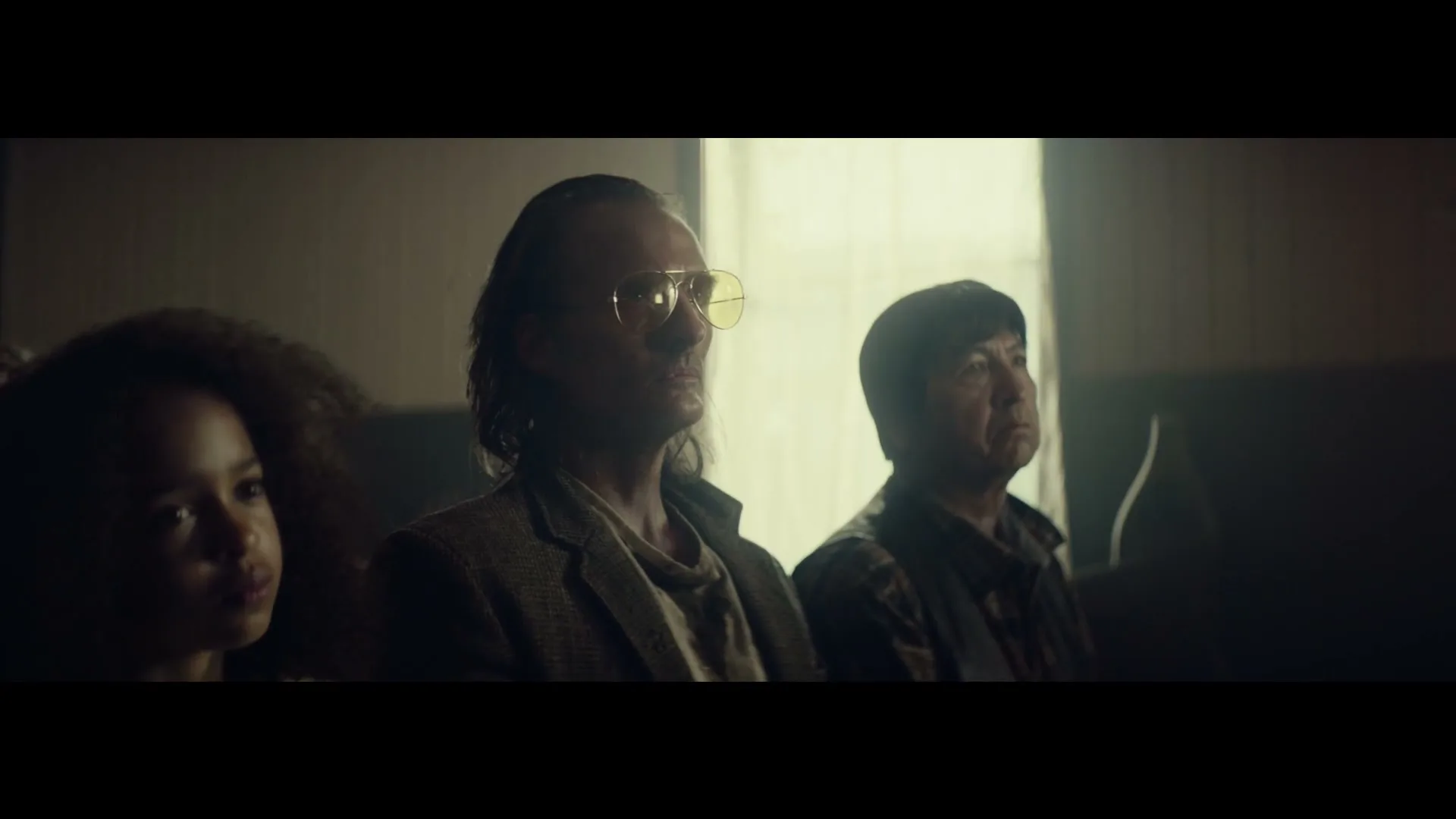 A young girl, a man in yellow glasses, and another man sit in a dimly lit room, looking up intently.
