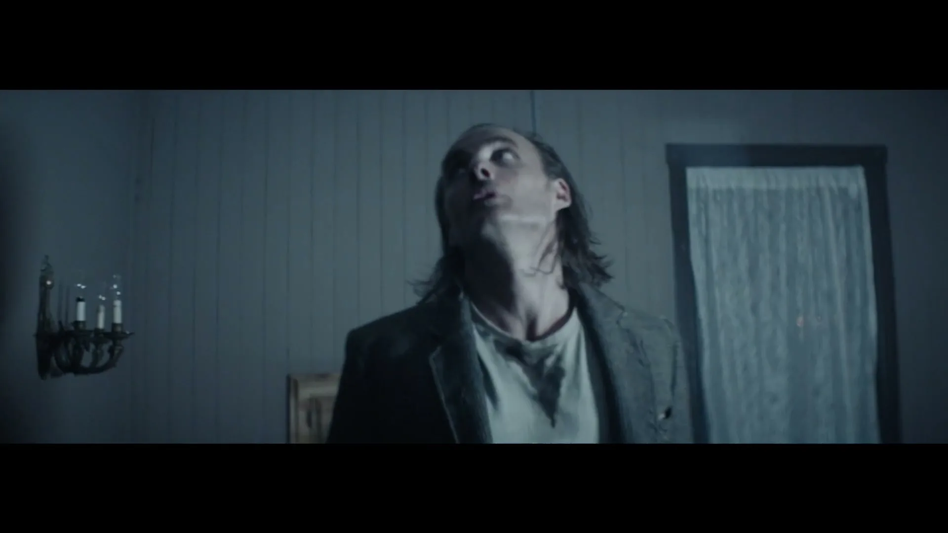 A man with long hair and a grey jacket looks up in a dimly lit room, appearing distressed.
