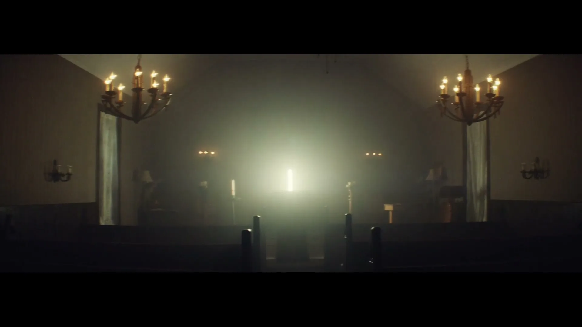 A wide shot of a dimly lit church interior with two chandeliers and a bright light source at the altar, creating a mysterious atmosphere.