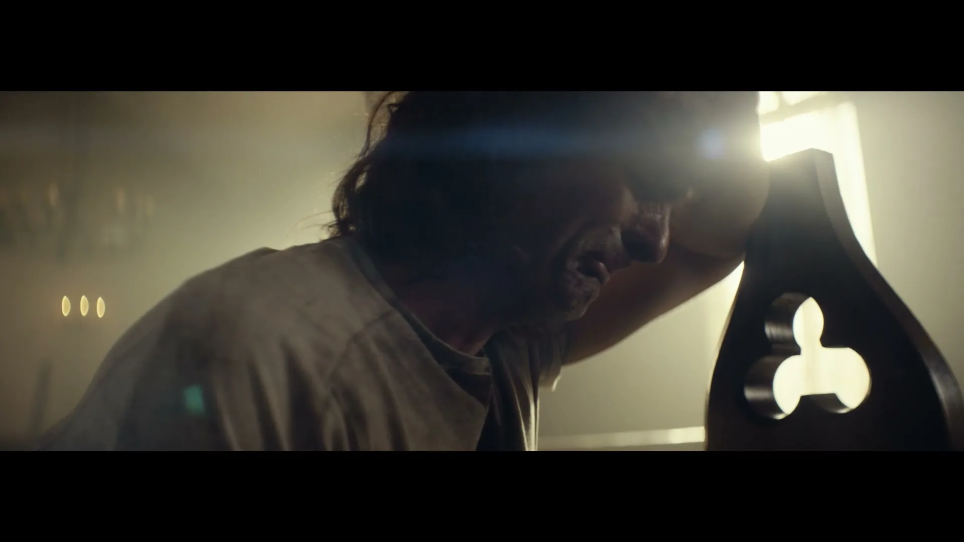 A man with long dark hair and a dirty t-shirt leans against a wooden pew in a smoky, dimly lit church, looking distressed.