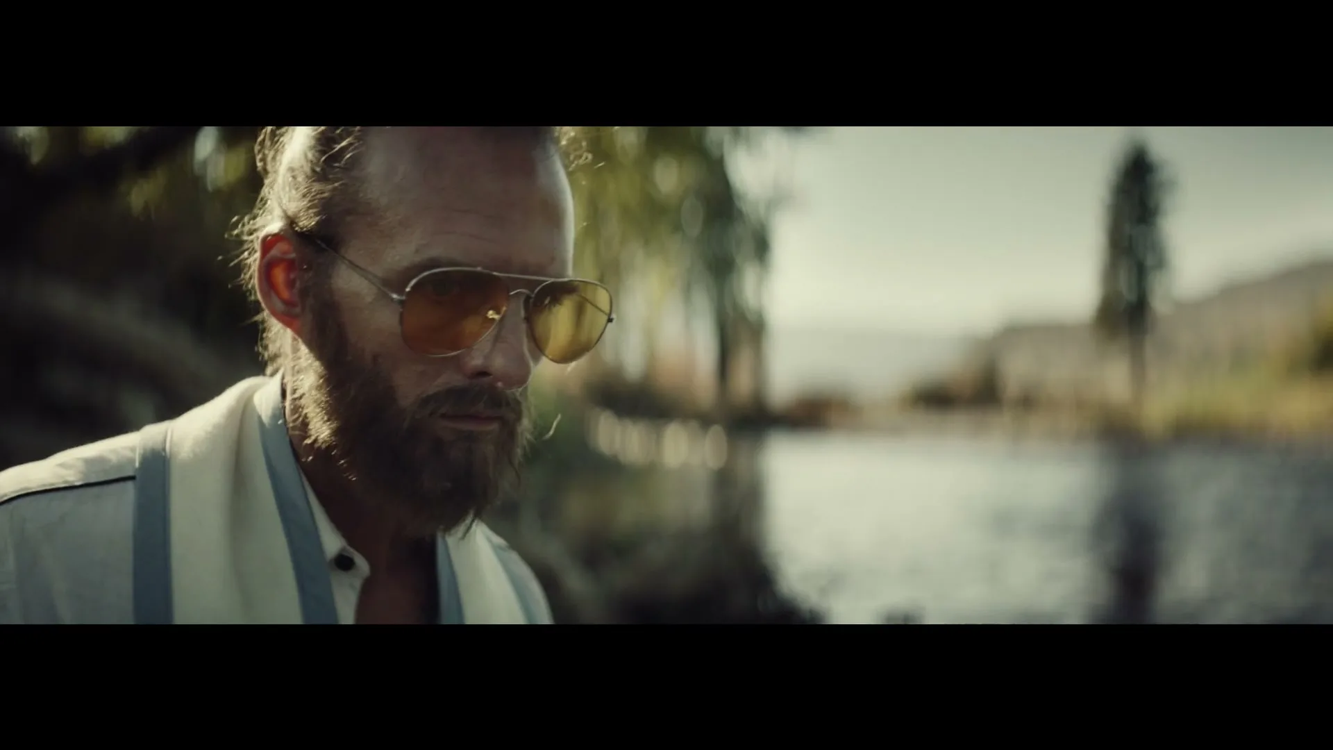 A bearded man in sunglasses and a striped shirt looks down, then up at the camera, by a body of water outdoors.