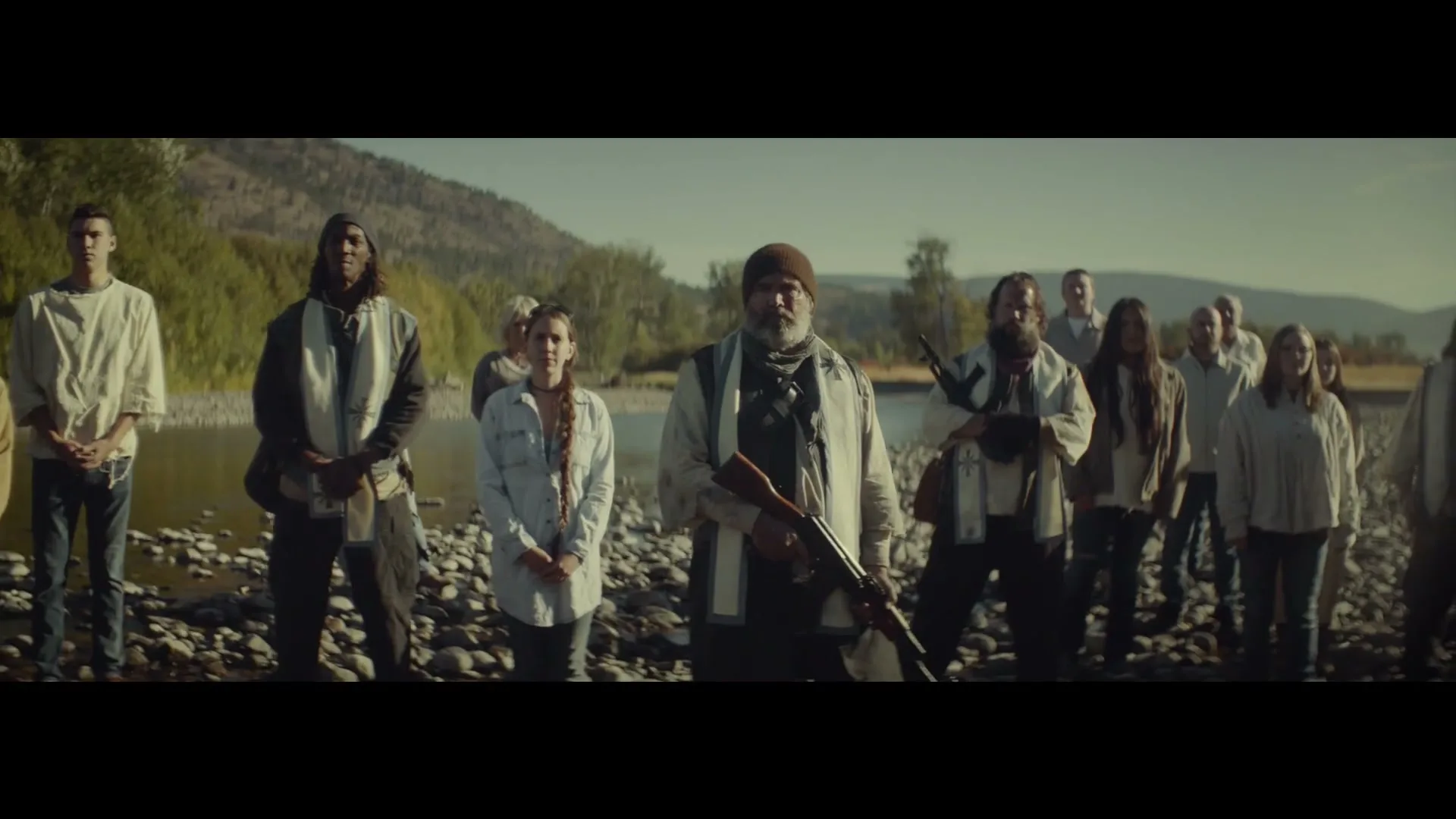 A group of people, some armed, stand on a rocky riverbank under a clear sky, looking directly at the camera.