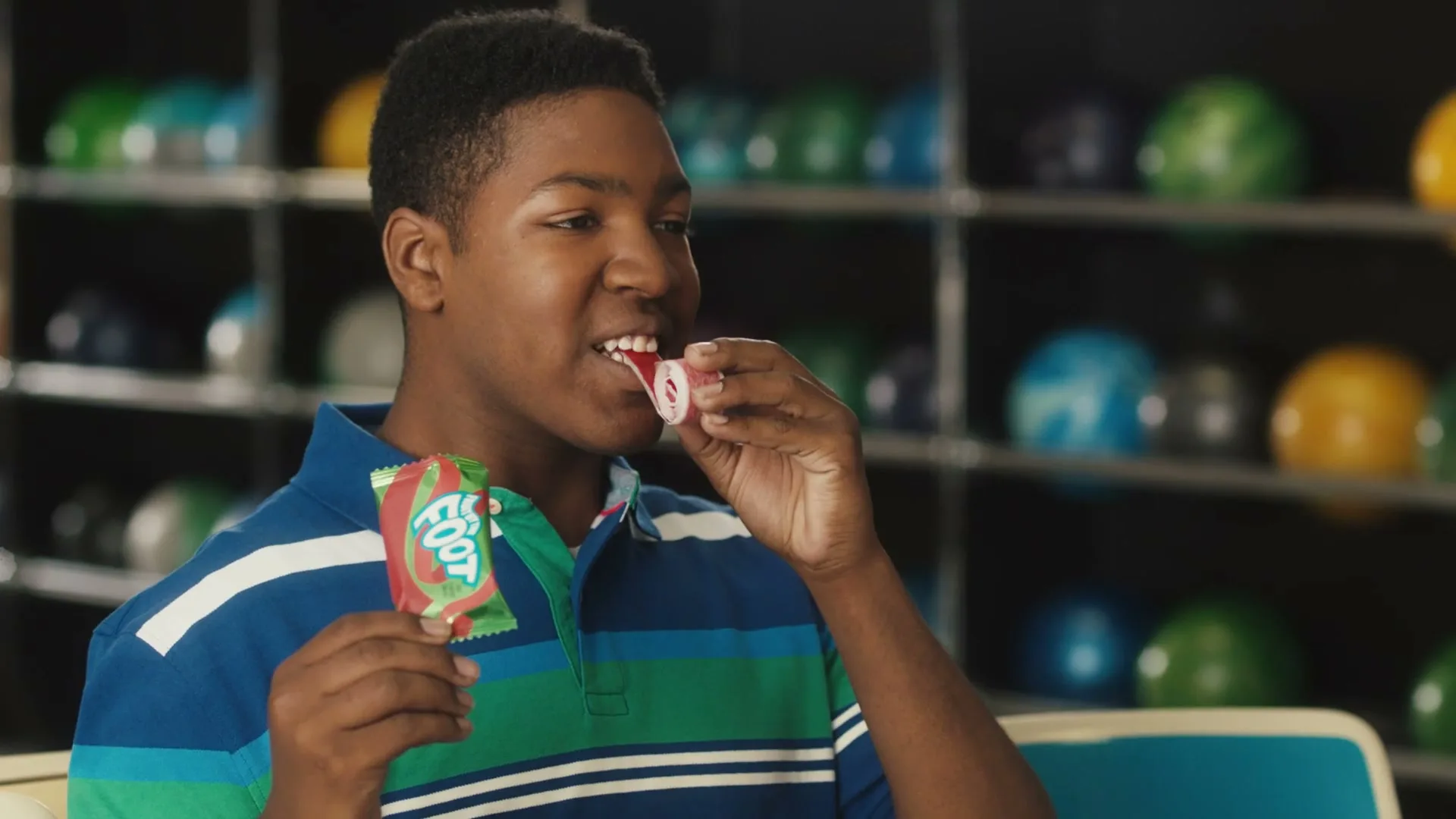 A young Black man in a blue and green polo shirt unwraps and eats a Fruit by the Foot in a bowling alley.
