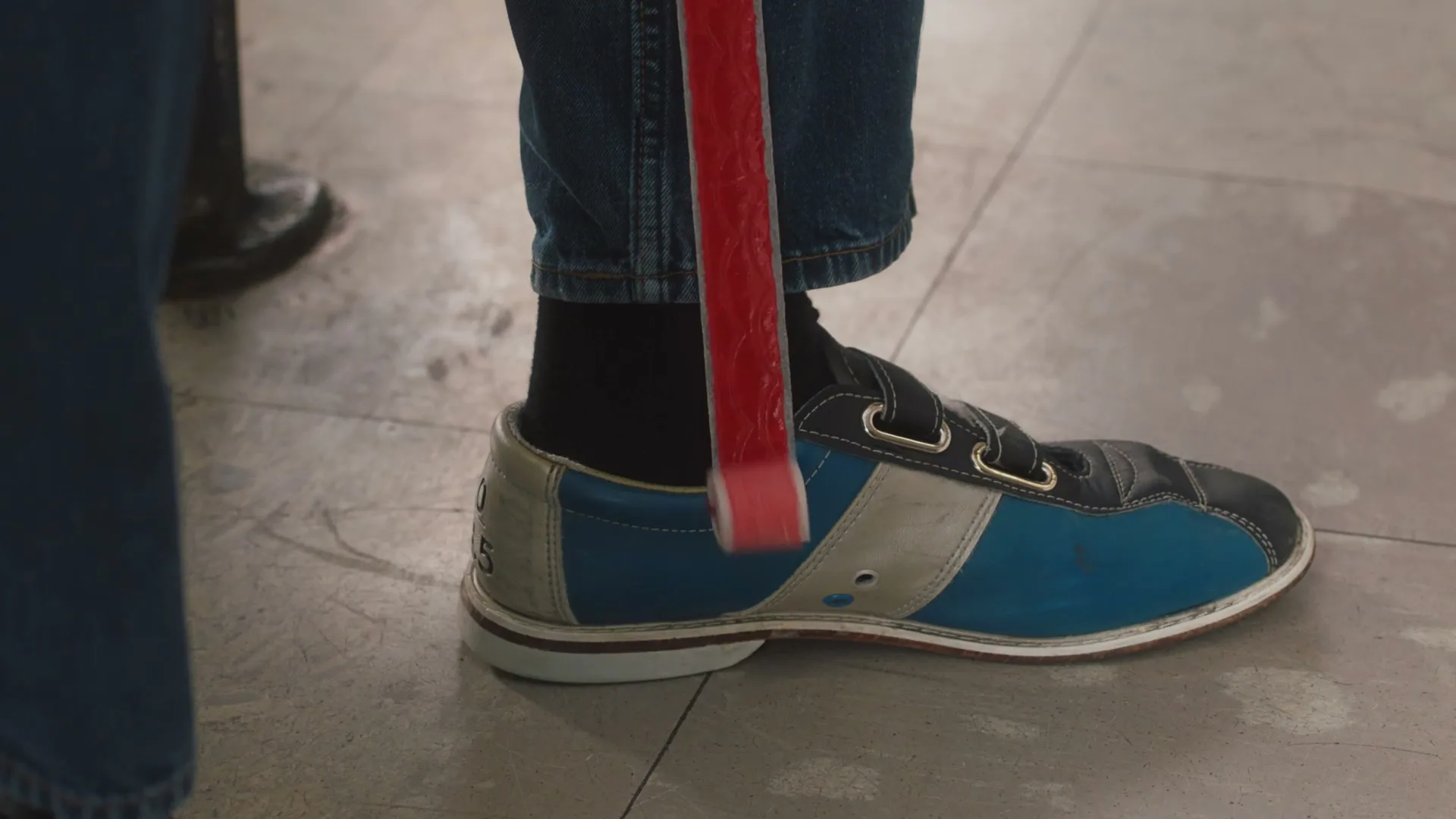 A person's foot in a blue and white bowling shoe is shown as a red Fruit by the Foot snack unrolls down their leg in a bowling alley.