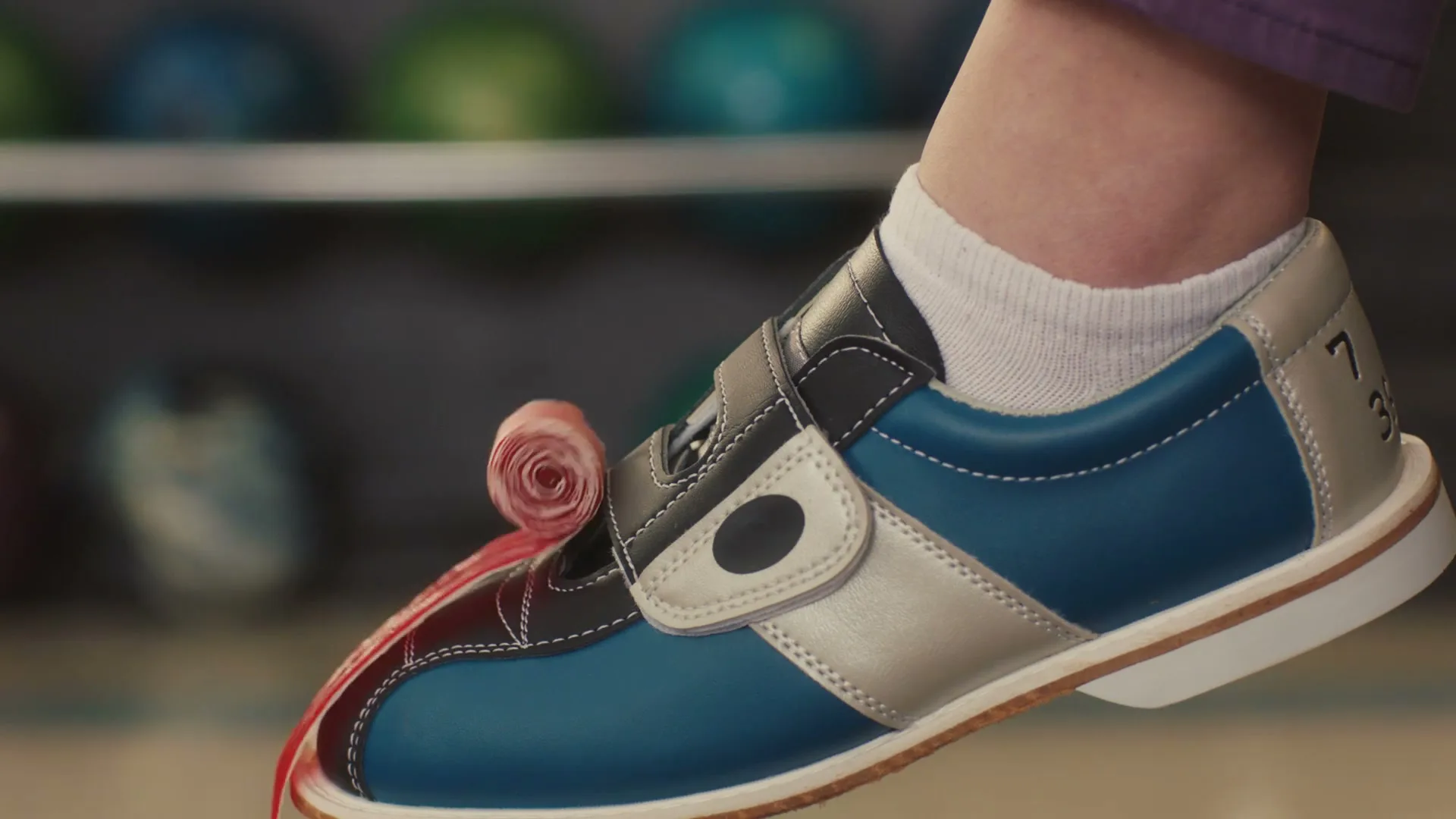 A person's foot in a blue and white bowling shoe is shown with a Fruit by the Foot candy unrolling from the toe, in a bowling alley.