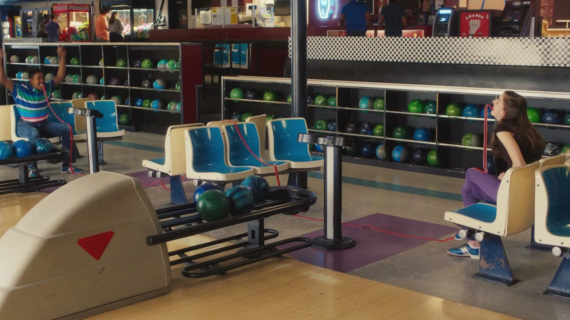 A young man and woman sit on opposite sides of a bowling alley, the man cheering with his arms up while the woman looks up with a Fruit by the Foot hanging from her mouth.