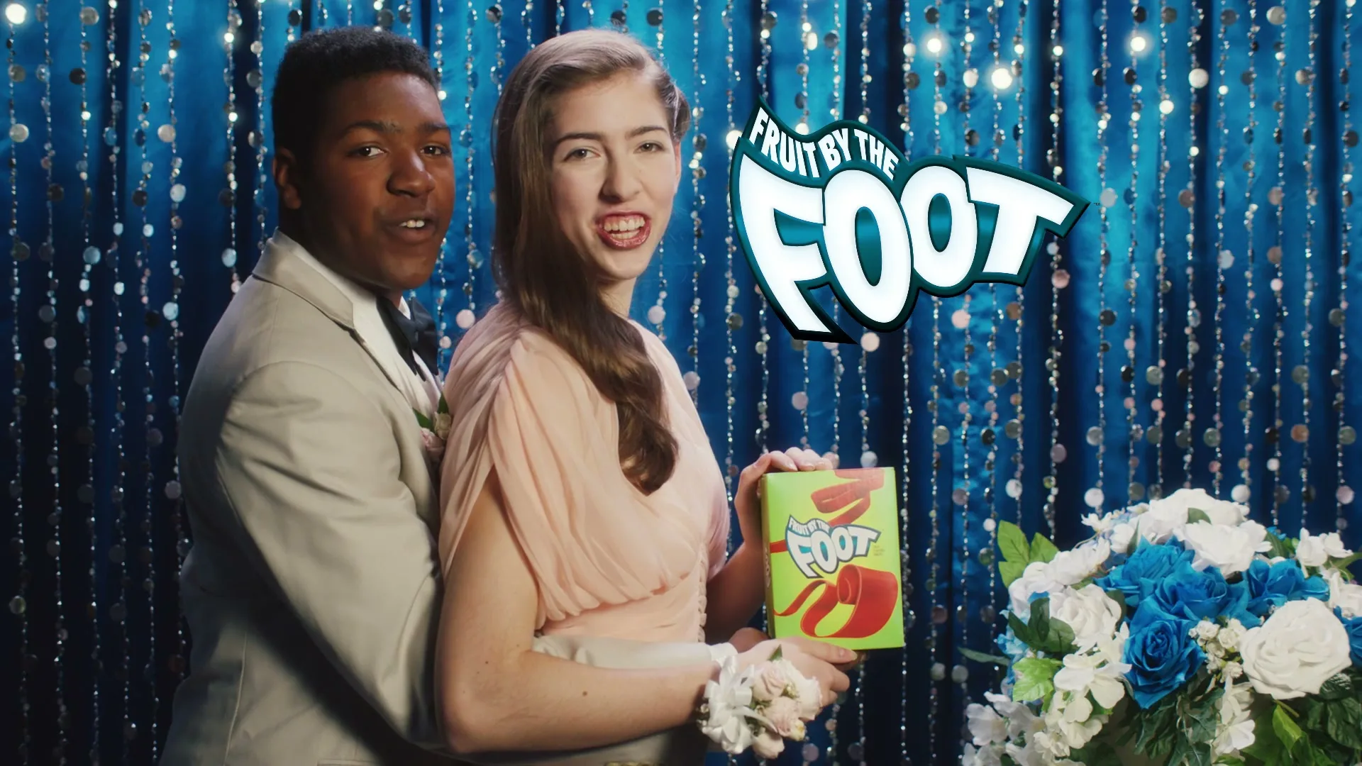 A young couple, dressed for prom, stands in front of a sparkly blue curtain, holding a Fruit by the Foot box and smiling at the camera.