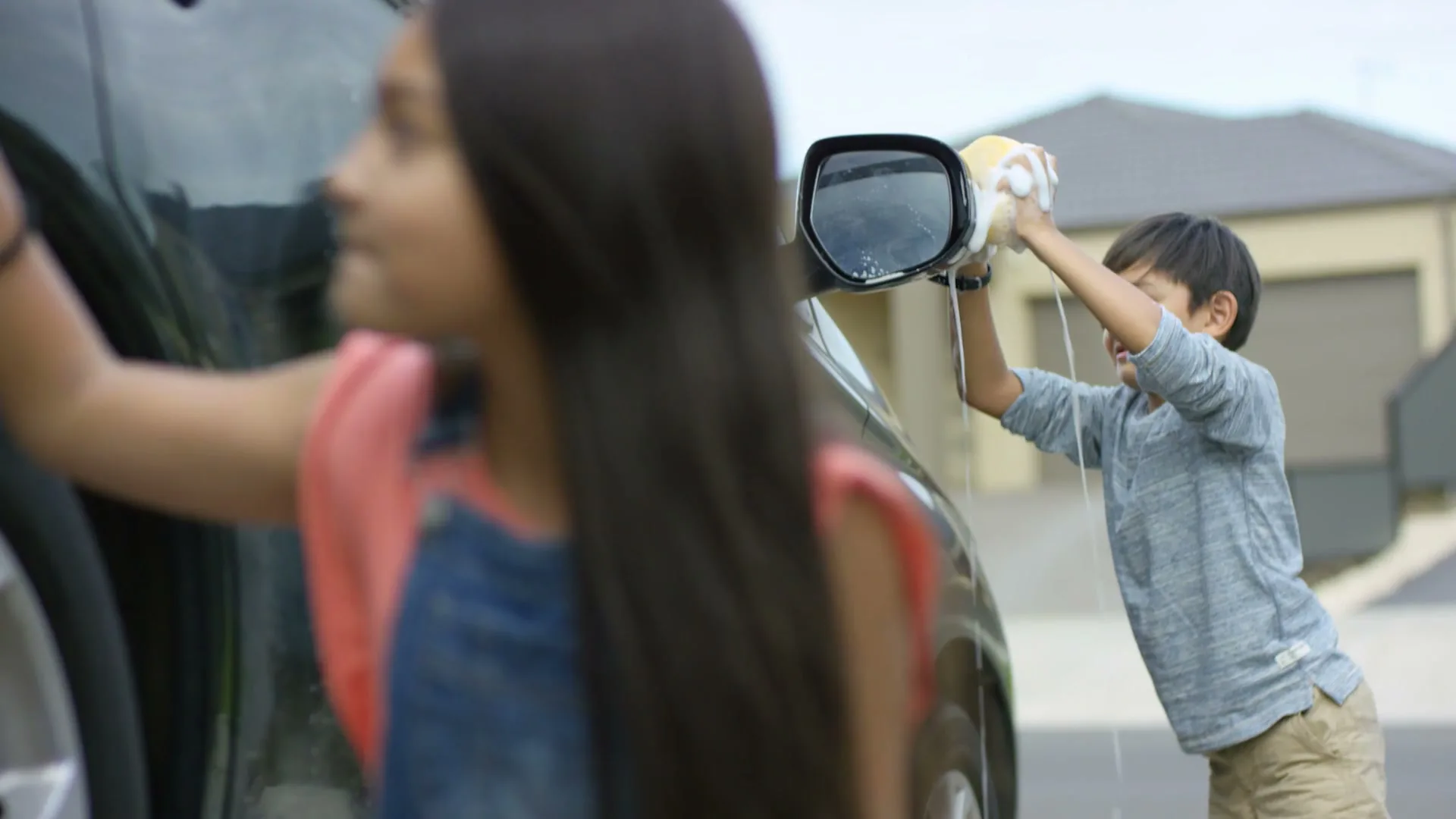 A young girl in denim overalls and a boy in a grey shirt wash a dark car in a suburban driveway on a bright day.