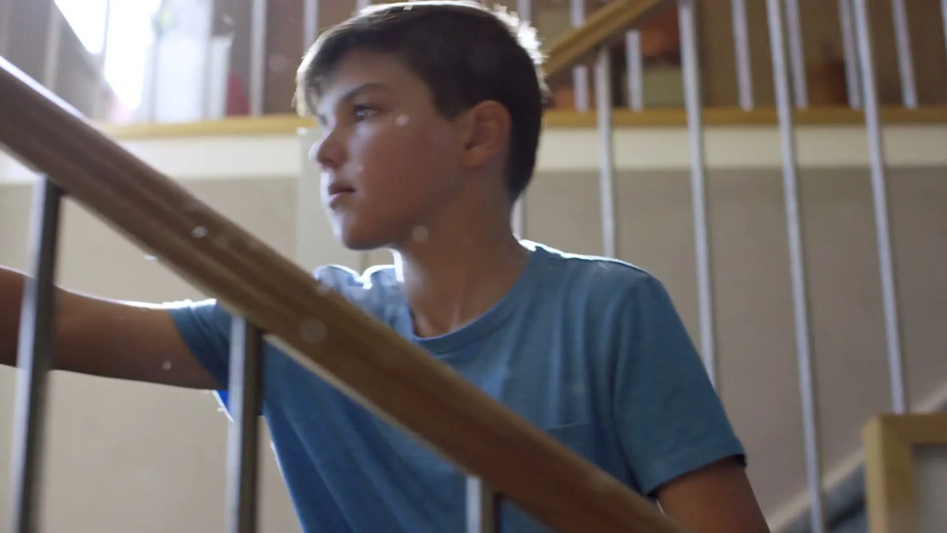 A young boy in a blue t-shirt dusts a wooden banister with a feather duster, then looks up as a girl follows him up the stairs in a bright house.