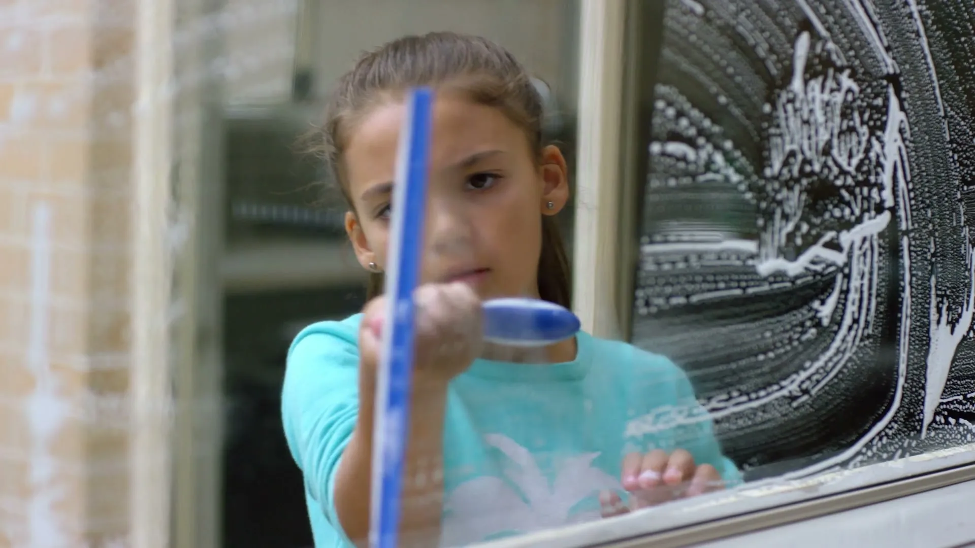A young girl with brown hair in a ponytail, wearing a teal long-sleeved shirt, cleans a window with a blue squeegee, looking focused.