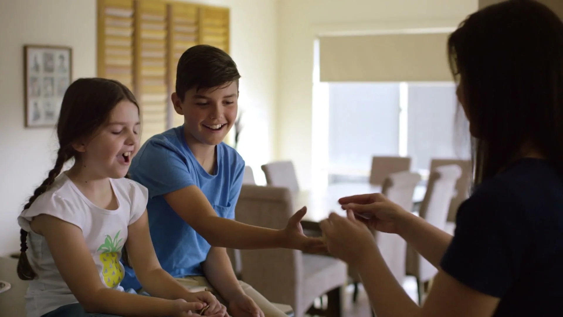 A mother gives pocket money to her two children, a boy and a girl, who are sitting at a table in a bright, modern home.