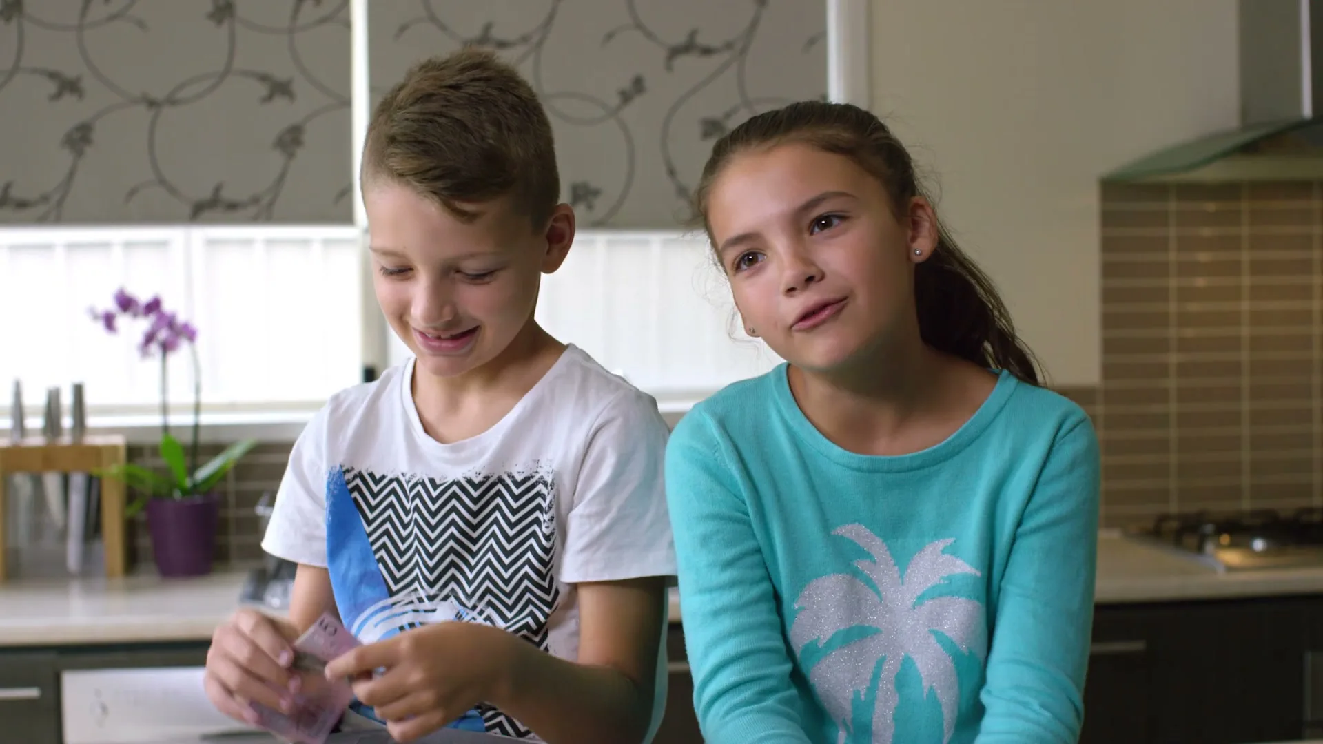 A young boy and girl sit side-by-side in a kitchen, the boy holding up a five-dollar bill and the girl looking at him with a questioning expression.