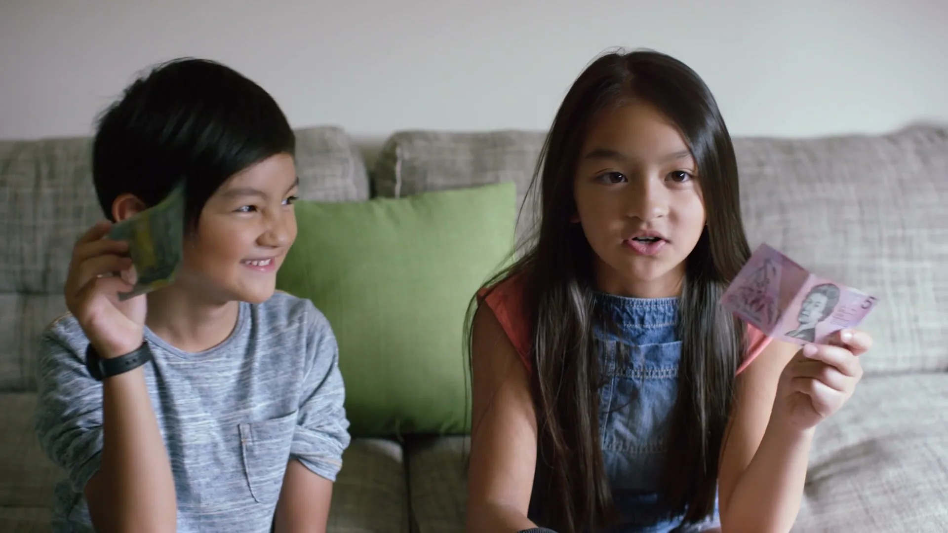 Two children, a boy and a girl, sit on a couch, holding up Australian banknotes and talking about them.