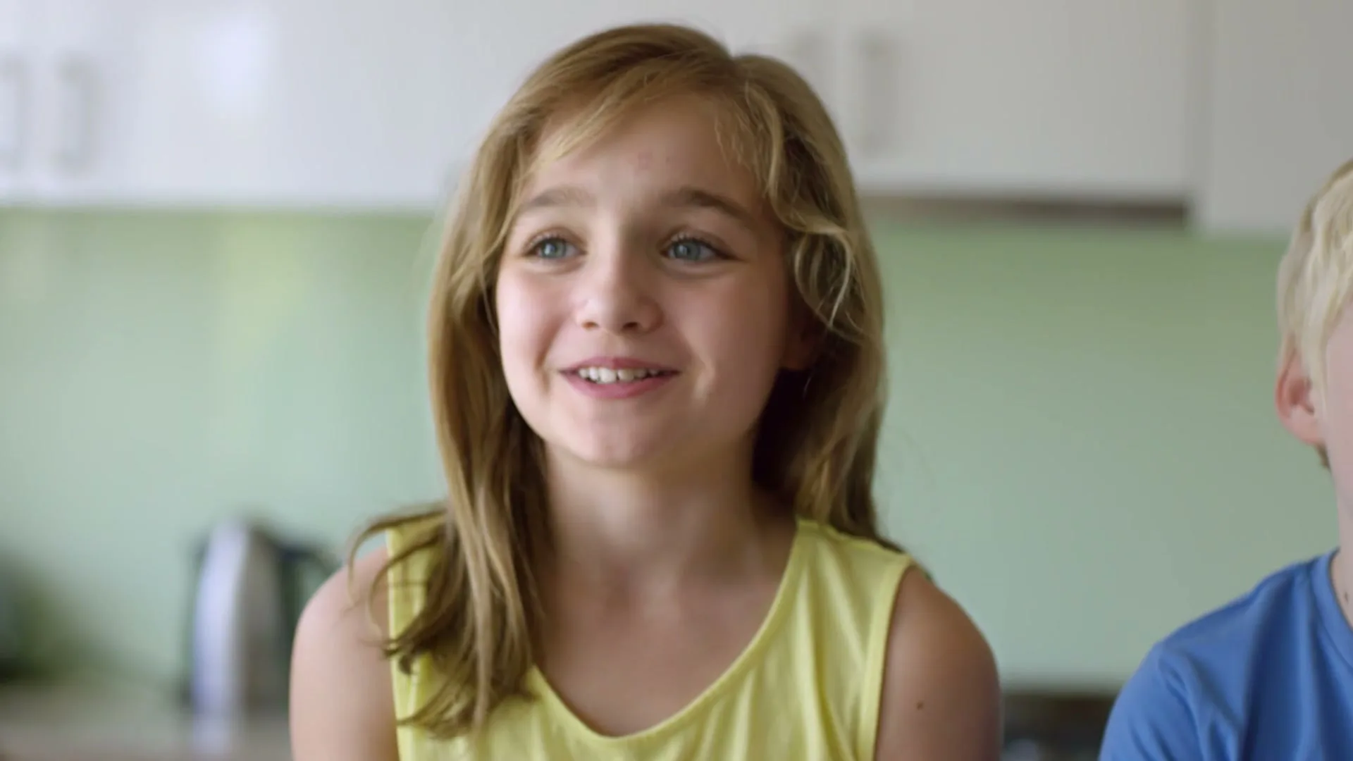 A young girl with blonde hair and a yellow top speaks directly to the camera, smiling and gesturing in a bright kitchen.