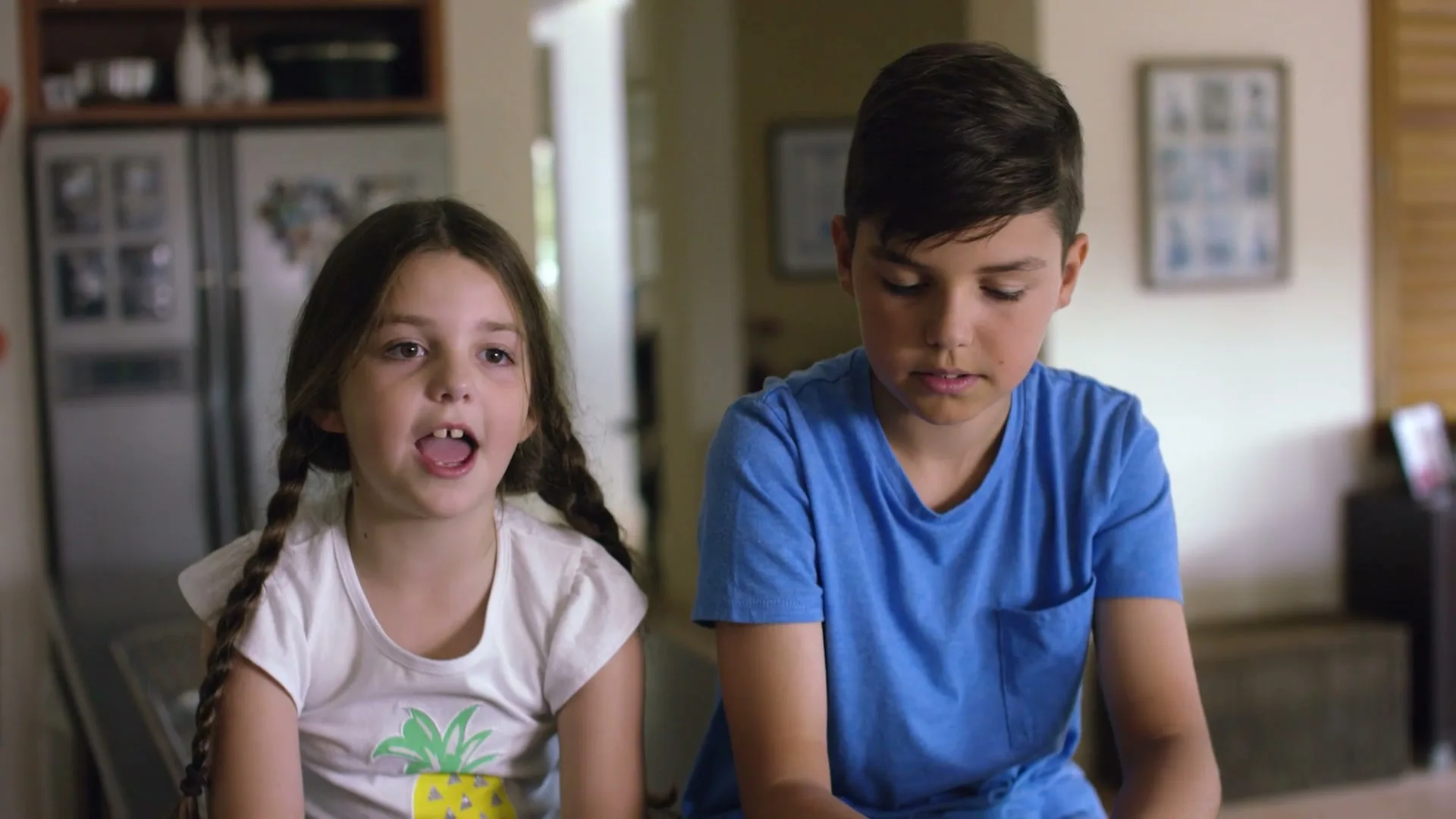 A young girl with pigtails and a boy in a blue shirt sit side-by-side, looking forward and talking in a brightly lit home interior.