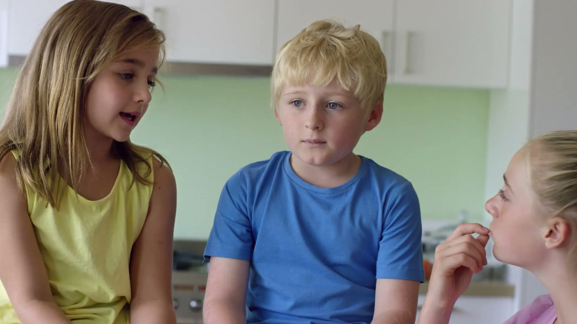 Three children, two girls and one boy, sit in a kitchen, talking amongst themselves about fairness.