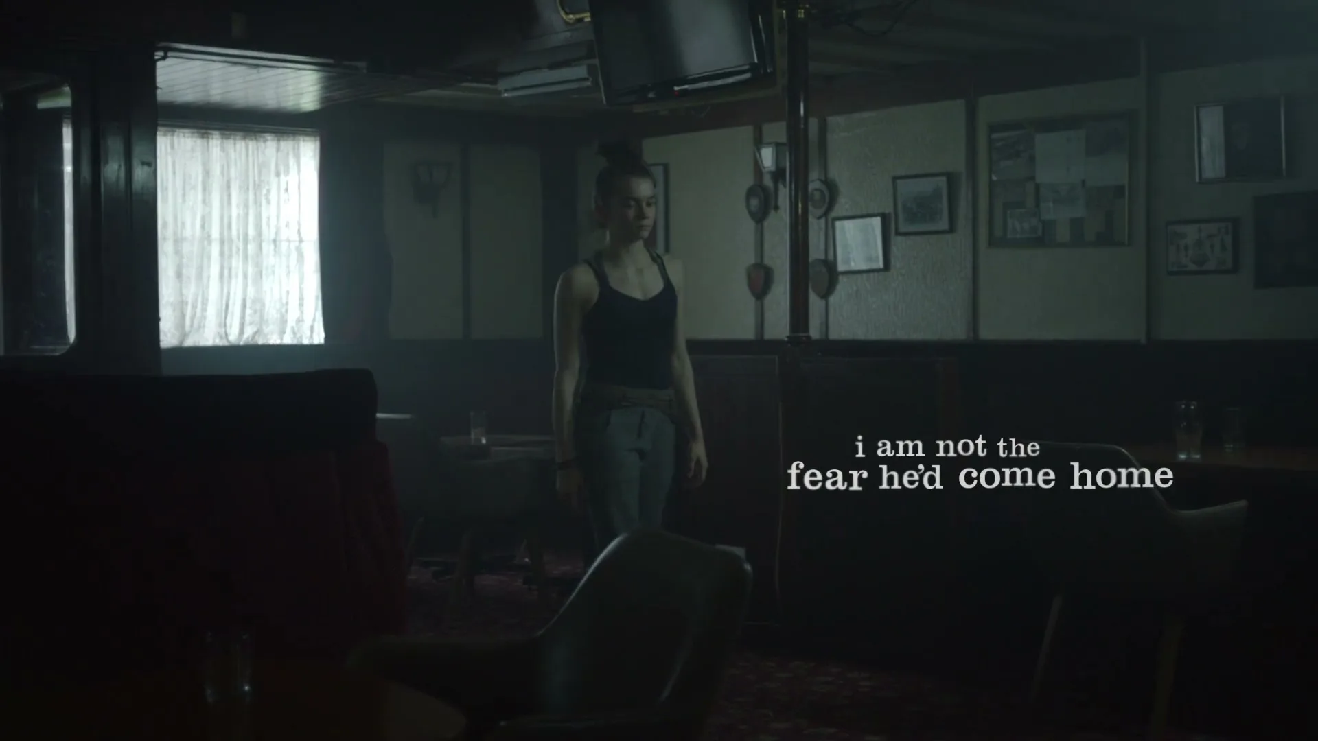 A young female gymnast with her hair in a bun practices her routine in a dimly lit, empty bar or restaurant, performing handstands and cartwheels.