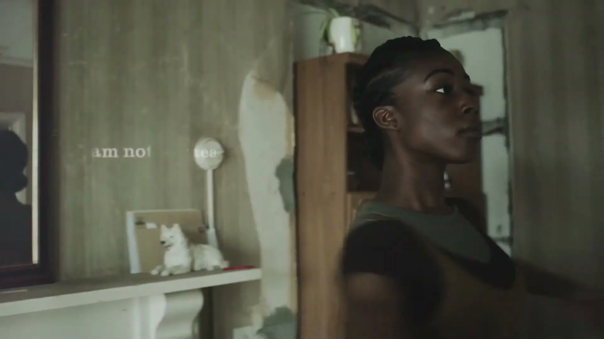 A young Black woman with braided hair, wearing a green top and brown overalls, looks around a dilapidated room with peeling wallpaper and a wooden cabinet.