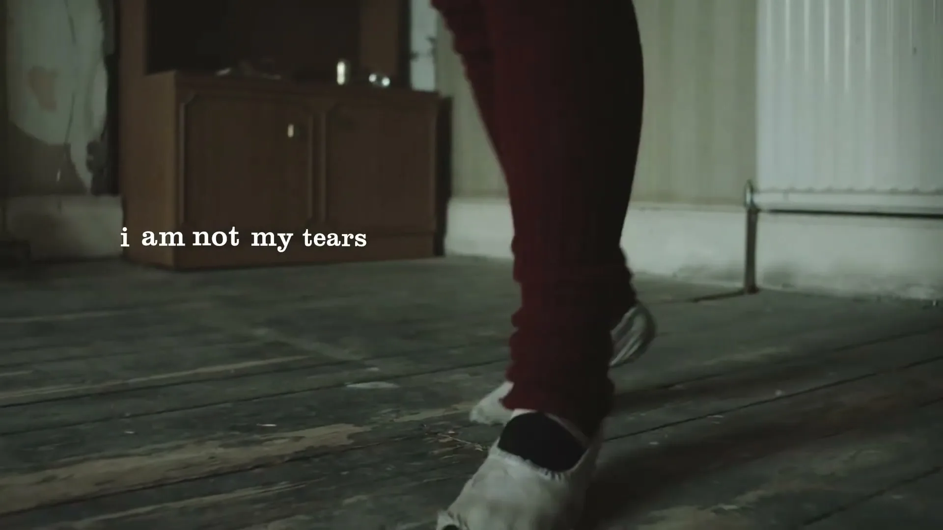 A young girl's feet in worn ballet slippers dance on a wooden floor in a dimly lit, empty room, with the text "i am not my tears" on screen.